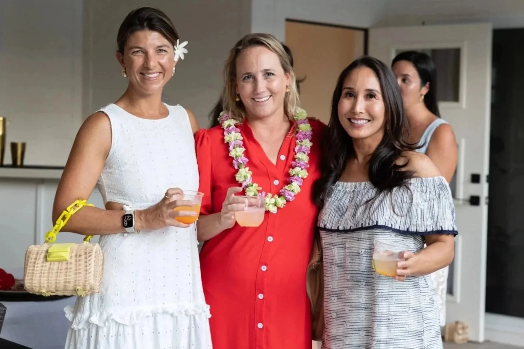 Four women at a social gathering, smiling and holding drinks, in a modern indoor setting.