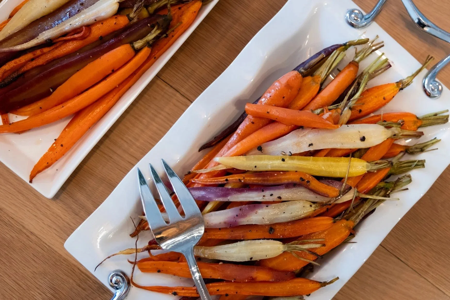Colorful roasted carrots on two white plates with a fork on one plate, placed on a wooden table.