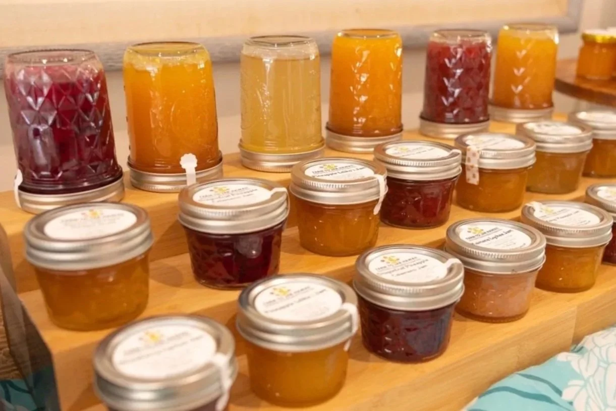 Jars and glasses of homemade jams and preserves on a wooden display at a market or shop.
