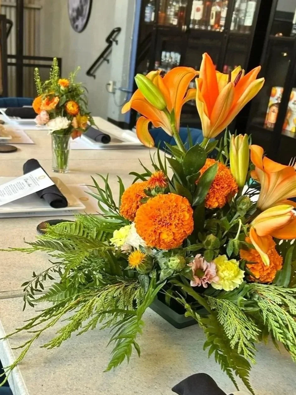 A vibrant floral arrangement featuring orange lilies, marigolds, and assorted greenery on a table in a room, with a smaller similar flower arrangement and a black cabinet in the background.