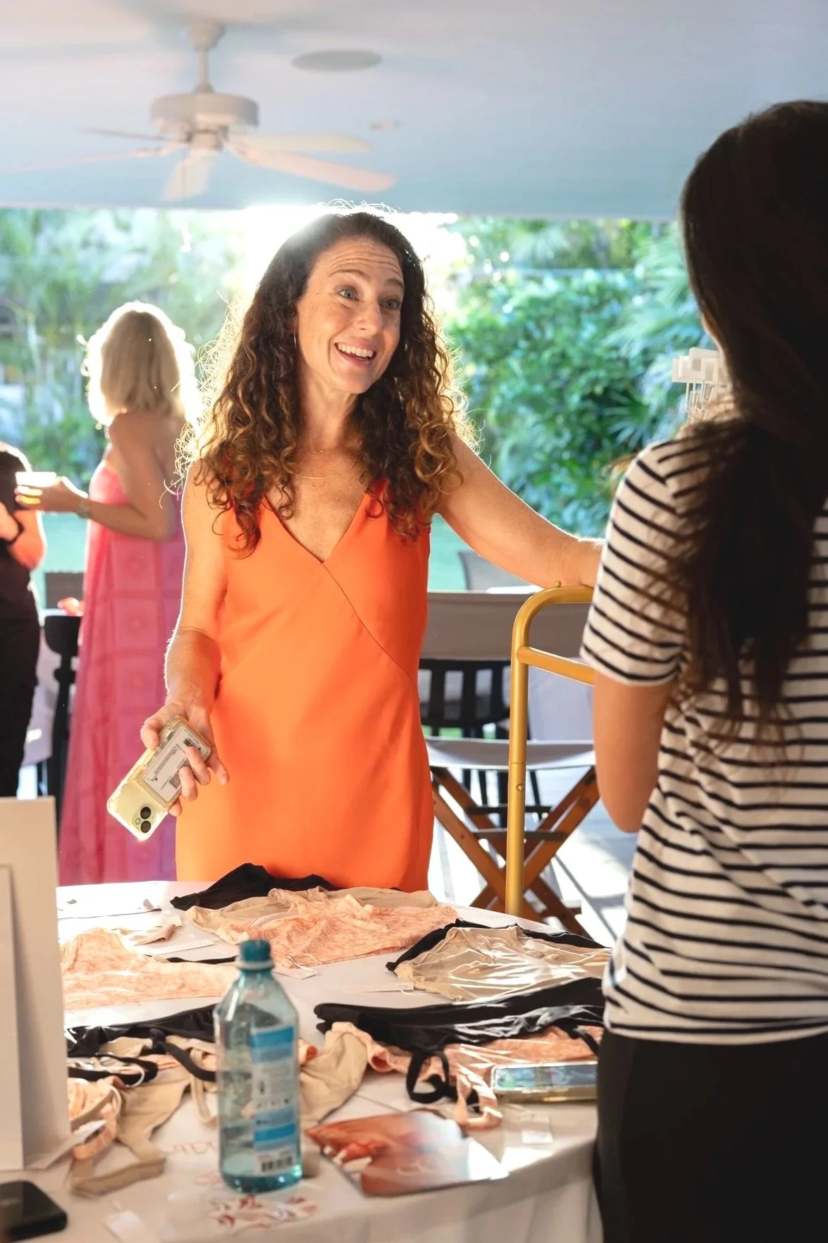 A woman in an orange dress at an outdoor event talking to another woman in a striped shirt, with a table of clothing and a water bottle in the foreground. Several people are in the background, and there is greenery outside.
