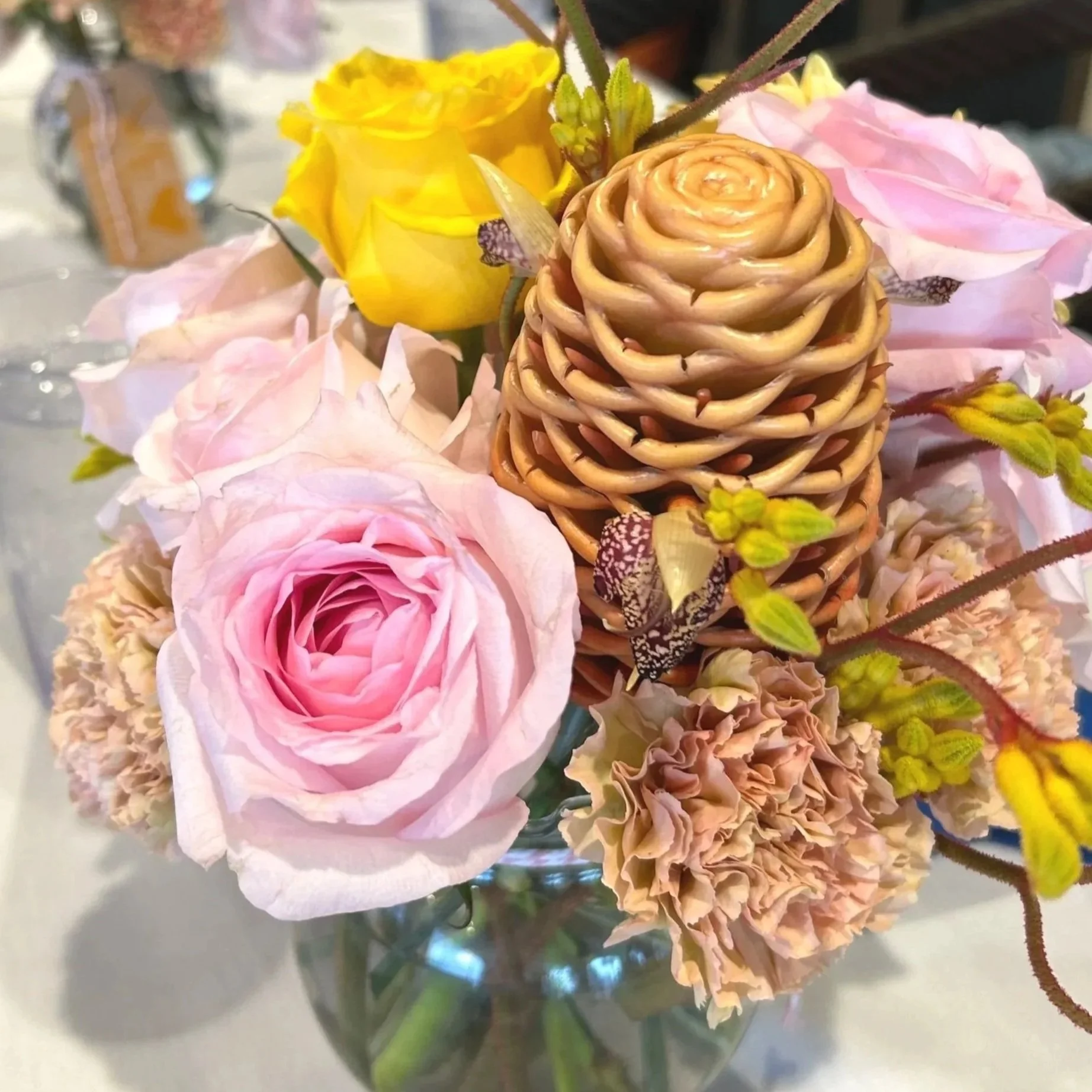 Close-up of a flower arrangement in a glass vase with pink roses, yellow flowers, and a pine cone.