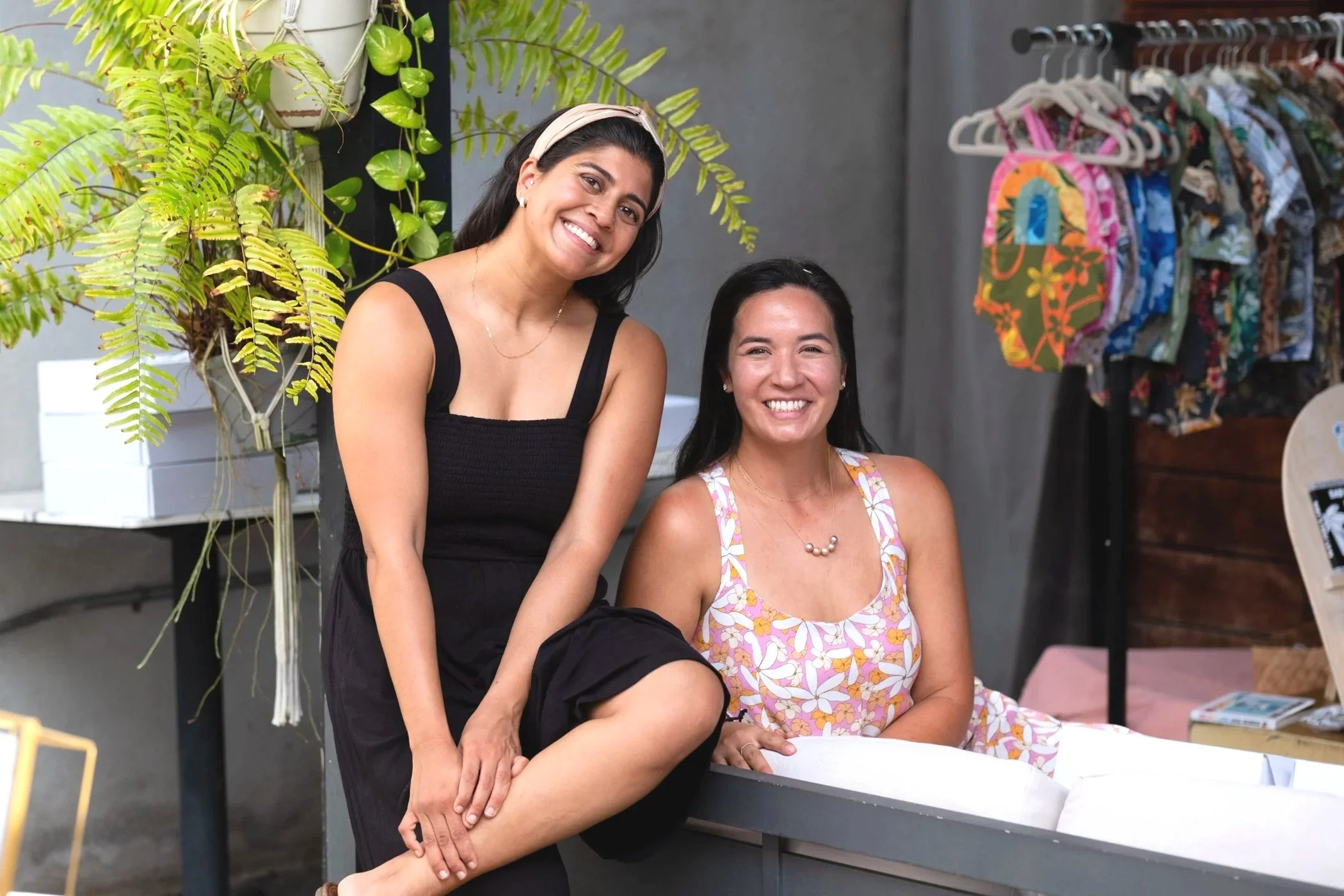 Two women smiling, one standing and one sitting, in a casual outdoor setting with clothing on hangers in the background and greenery nearby.