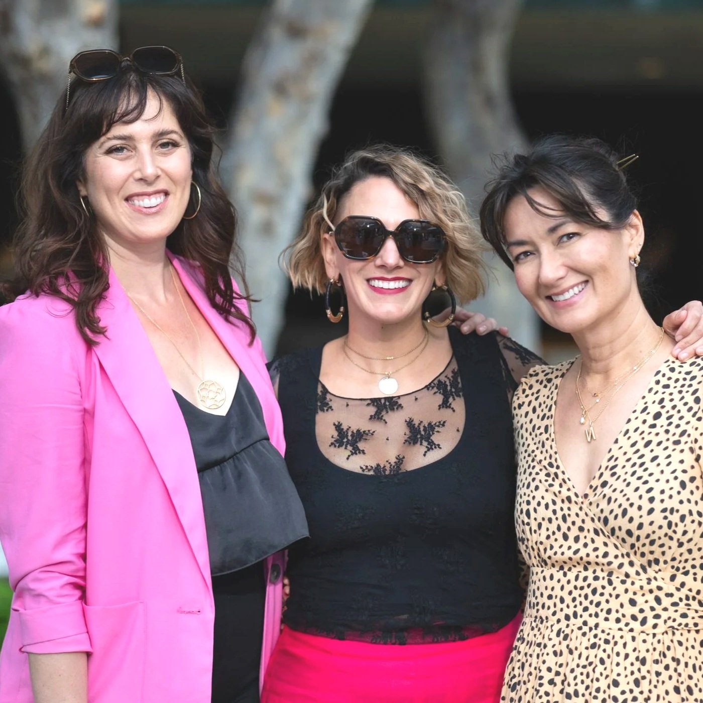 Three women smiling outdoors, one in a pink blazer, one in a black lace top with sunglasses, and one in a leopard print dress.