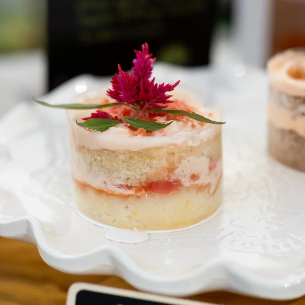 A small layered dessert in a clear cylindrical container, topped with pink flower and green leaves, placed on a white doily