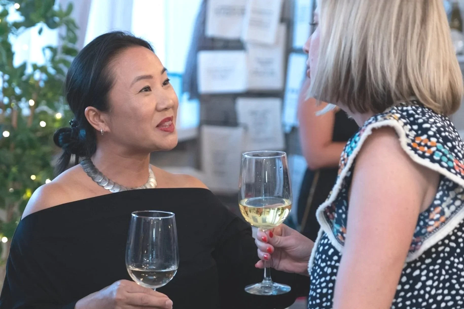 Two women engaging in conversation at a social gathering, each holding a glass of white wine. The woman on the left, with dark hair styled back and wearing a black off-shoulder dress with a silver necklace, smile warmly. The woman on the right, with 