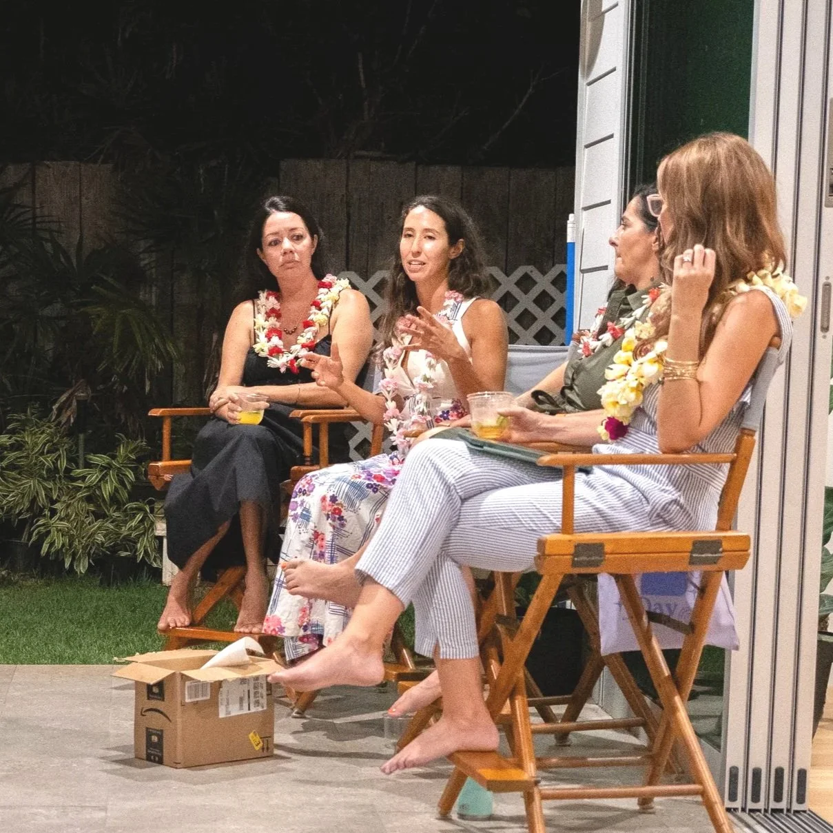 Four women wearing leis sit on wooden chairs outdoors at night, engaging in conversation with drinks in hand.
