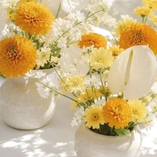 Two white vases with yellow, orange, and white flowers, including marigolds, daisies, and calla lilies, on a white surface.