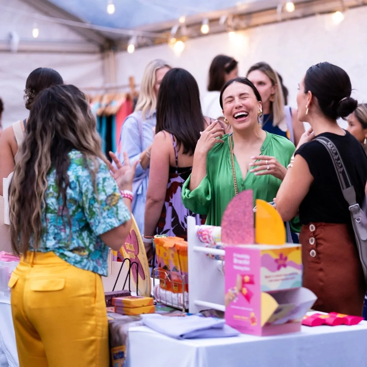 A woman in a green top laughs with a group of women at an outdoor craft fair or market, with various colorful products on display.