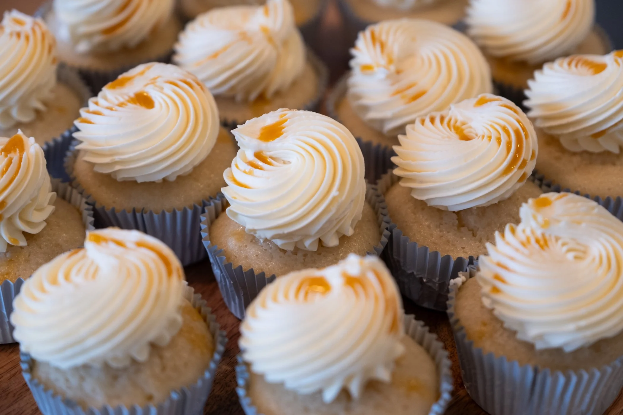 A close-up view of several cupcakes topped with swirls of white frosting and caramel drizzle, each in a gray cupcake liner.