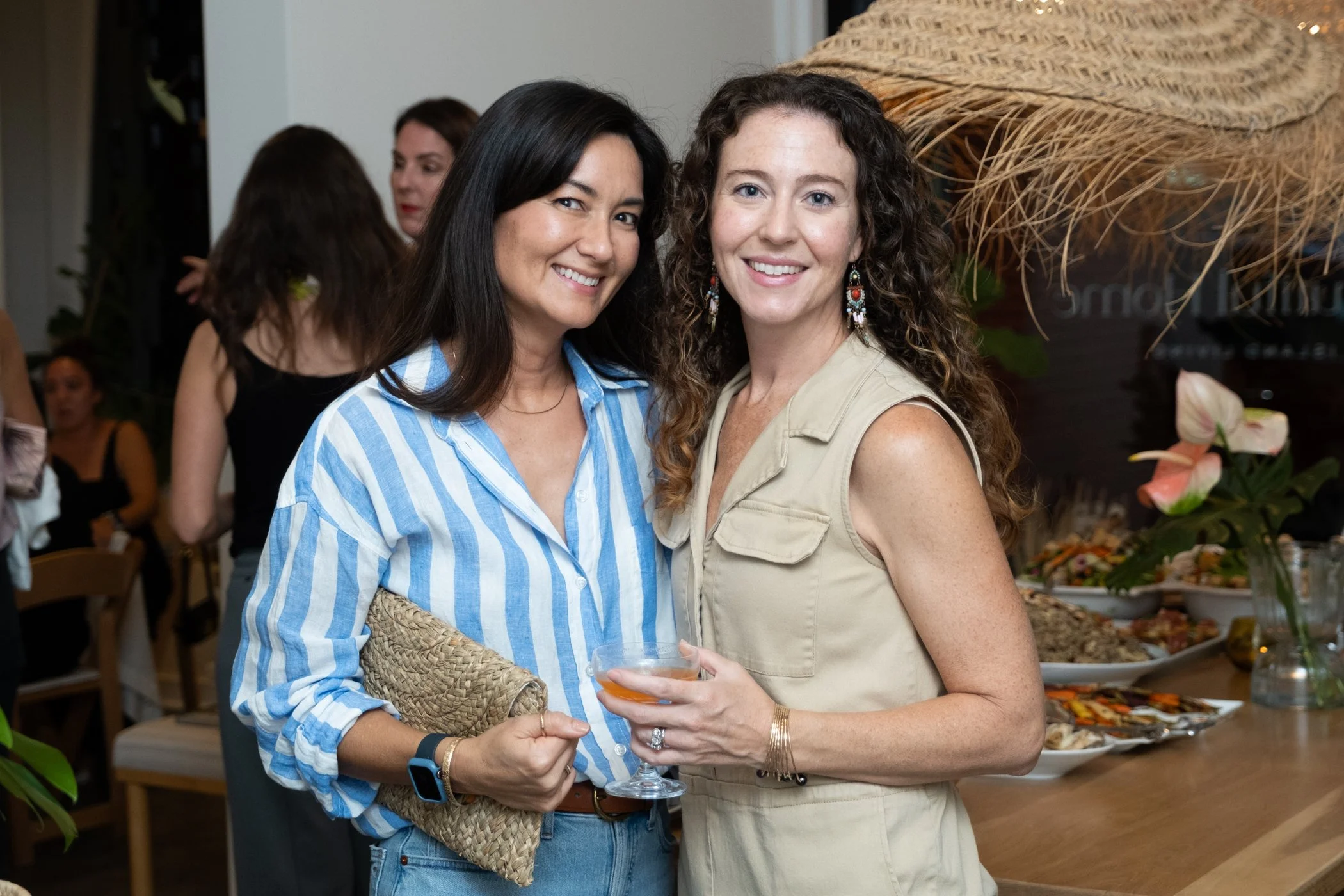 Two women smiling at a party, one with dark hair wearing a striped blue and white shirt and the other with curly hair wearing a beige dress, standing near a table with food and decorations.