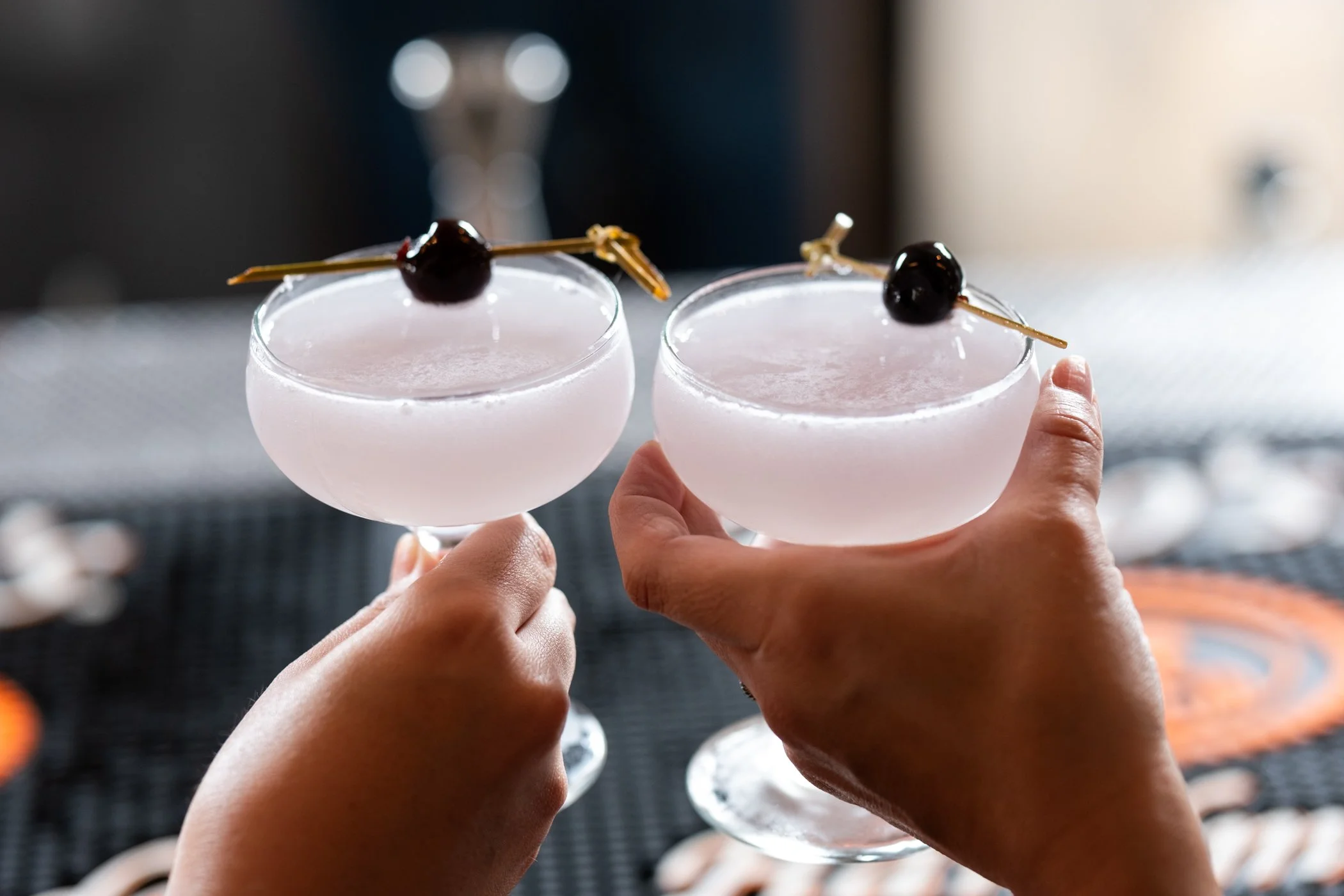 Two hands holding pink cocktail glasses topped with cherries and garnished with skewers, with a blurred background.