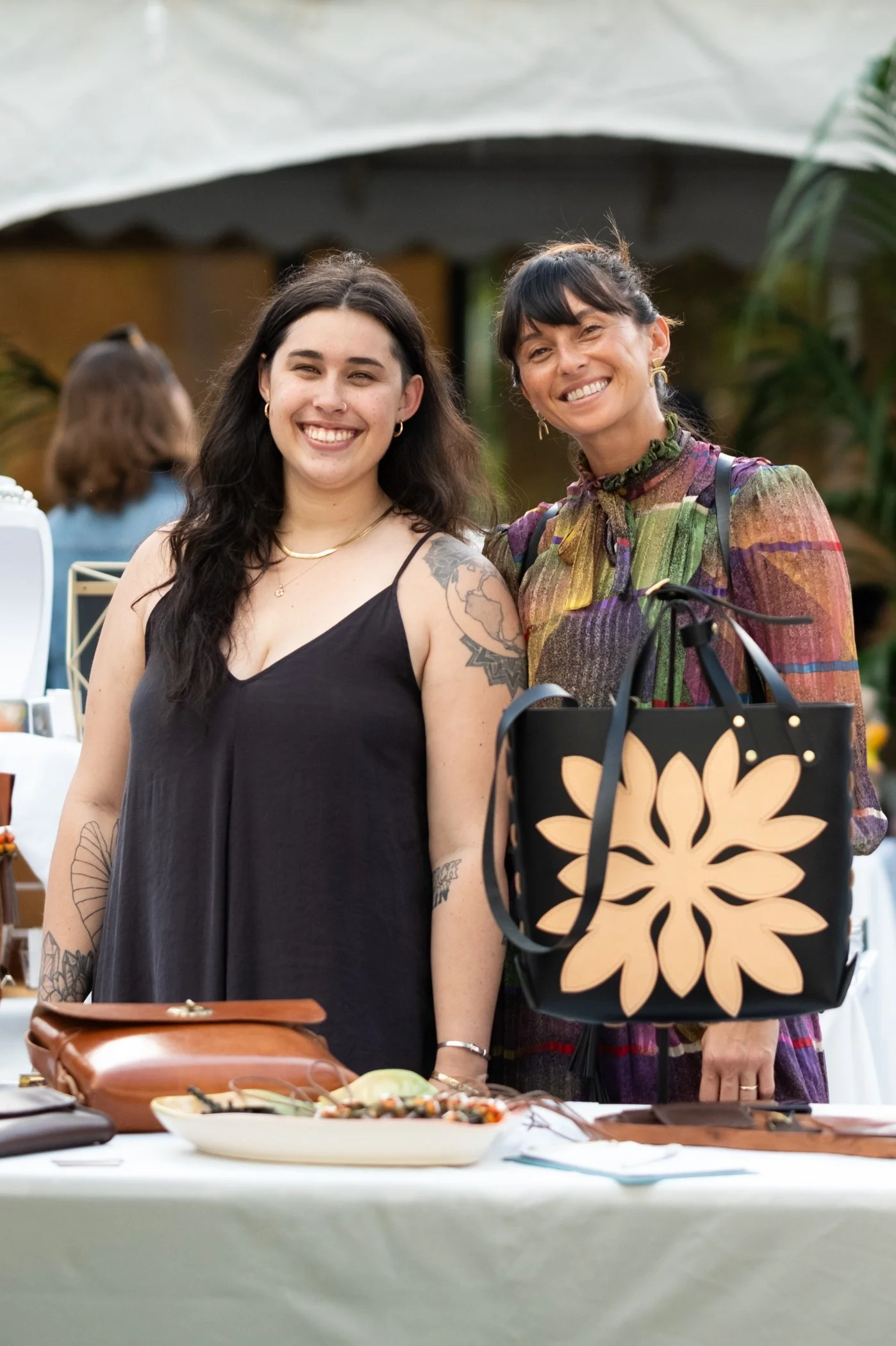 Two women standing behind a table at an outdoor event, smiling at the camera. One woman has dark hair, tattoos, and is wearing a black sleeveless dress, and the other woman has dark hair with bangs, earrings, and is wearing a colorful patterned dress
