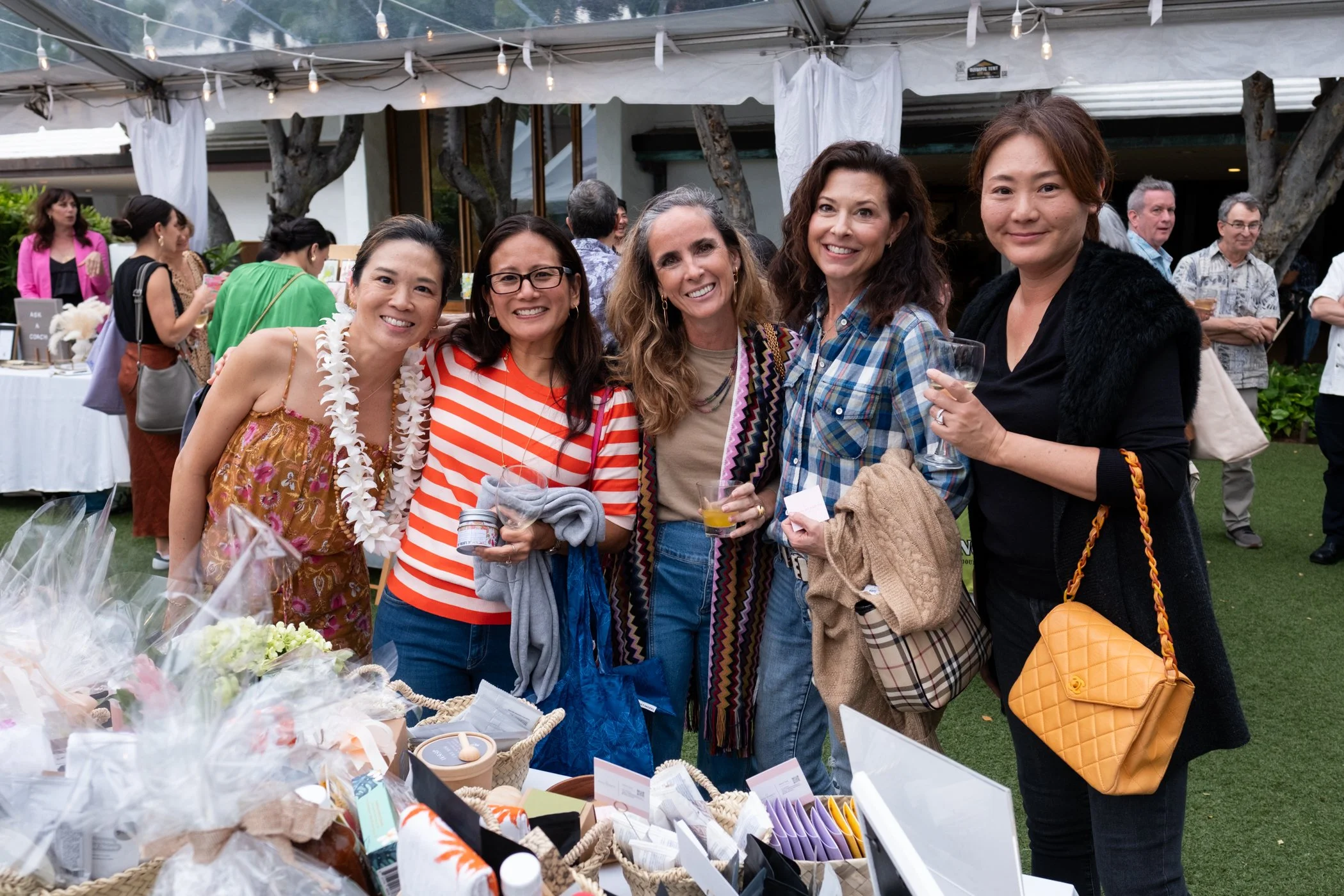 Group of five women smiling at a social event, standing outdoors under a tent with string lights. They are surrounded by various tables with items for sale or display, including flowers and cards.