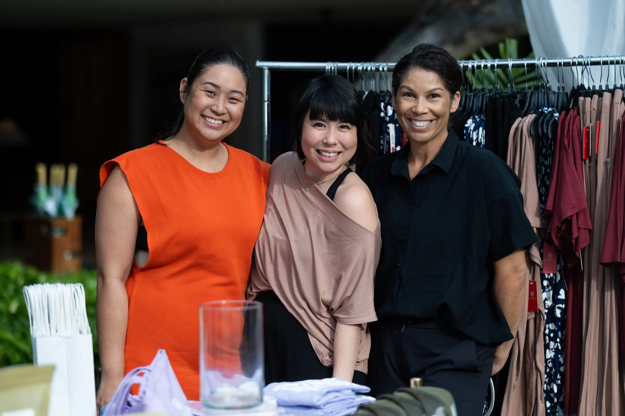 Three women smiling at the camera at an outdoor event with clothing racks in the background.