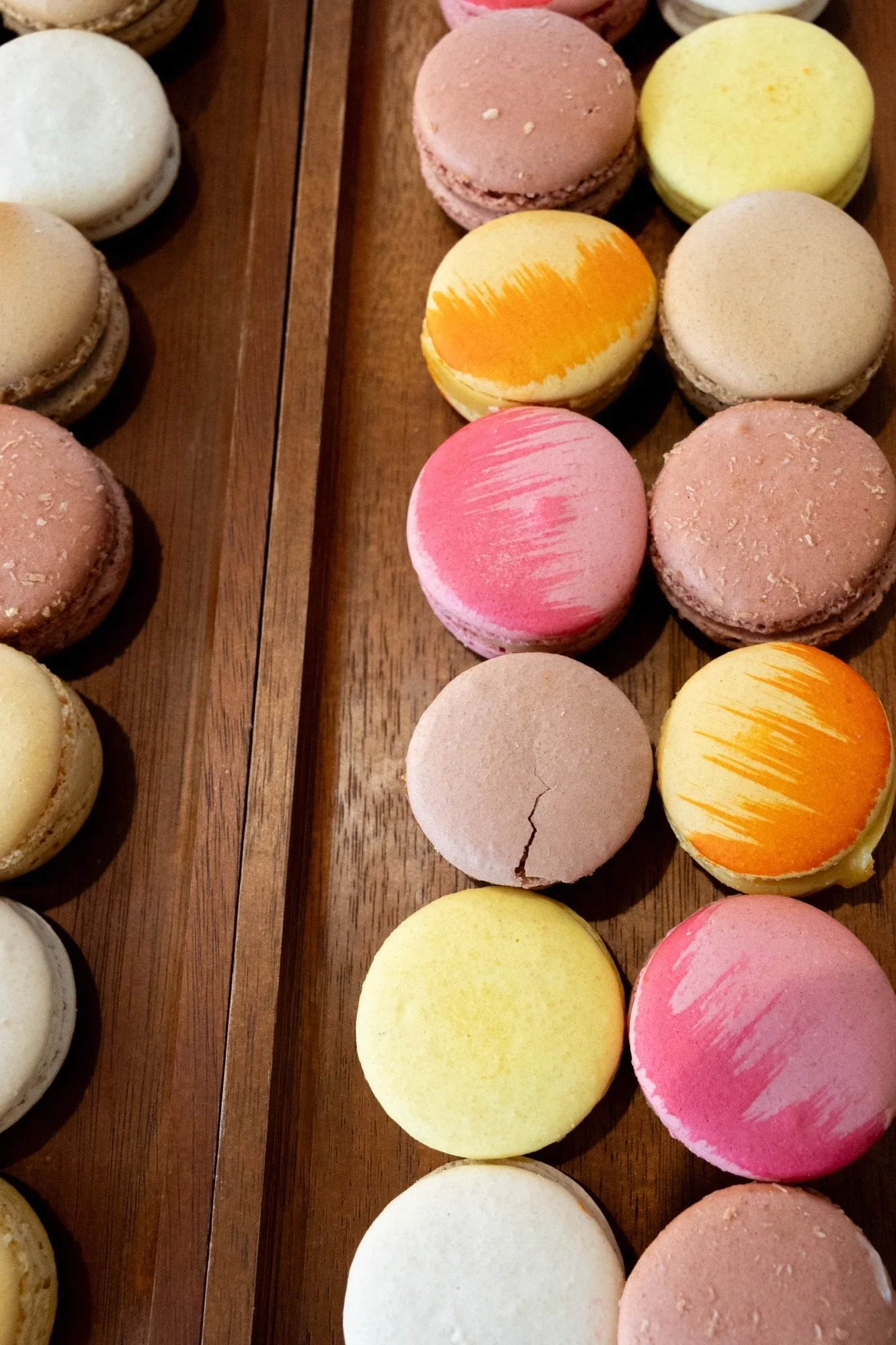 Colorful macarons arranged on a wooden surface in rows with various pastel shades including pink, yellow, white, and light brown.