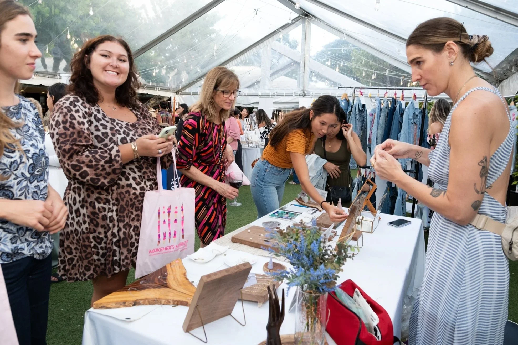 People browsing handmade jewelry at an outdoor market under a large transparent tent with string lights. Several women are looking at various jewelry pieces on display.