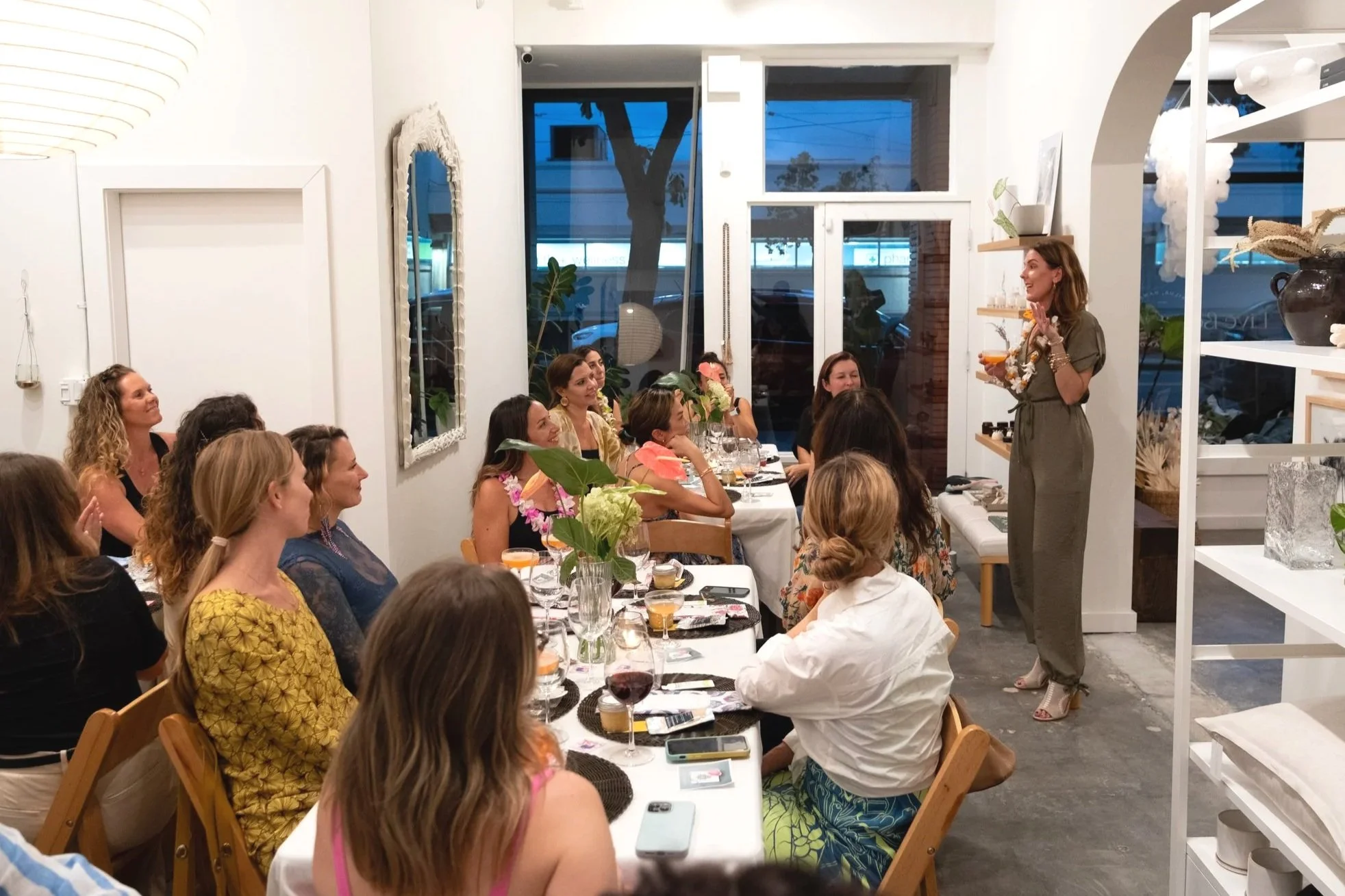 A woman giving a speech or presentation to a group of women seated at a long dining table in a well-lit, modern indoor space with large windows, indoor plants, and decorative items.