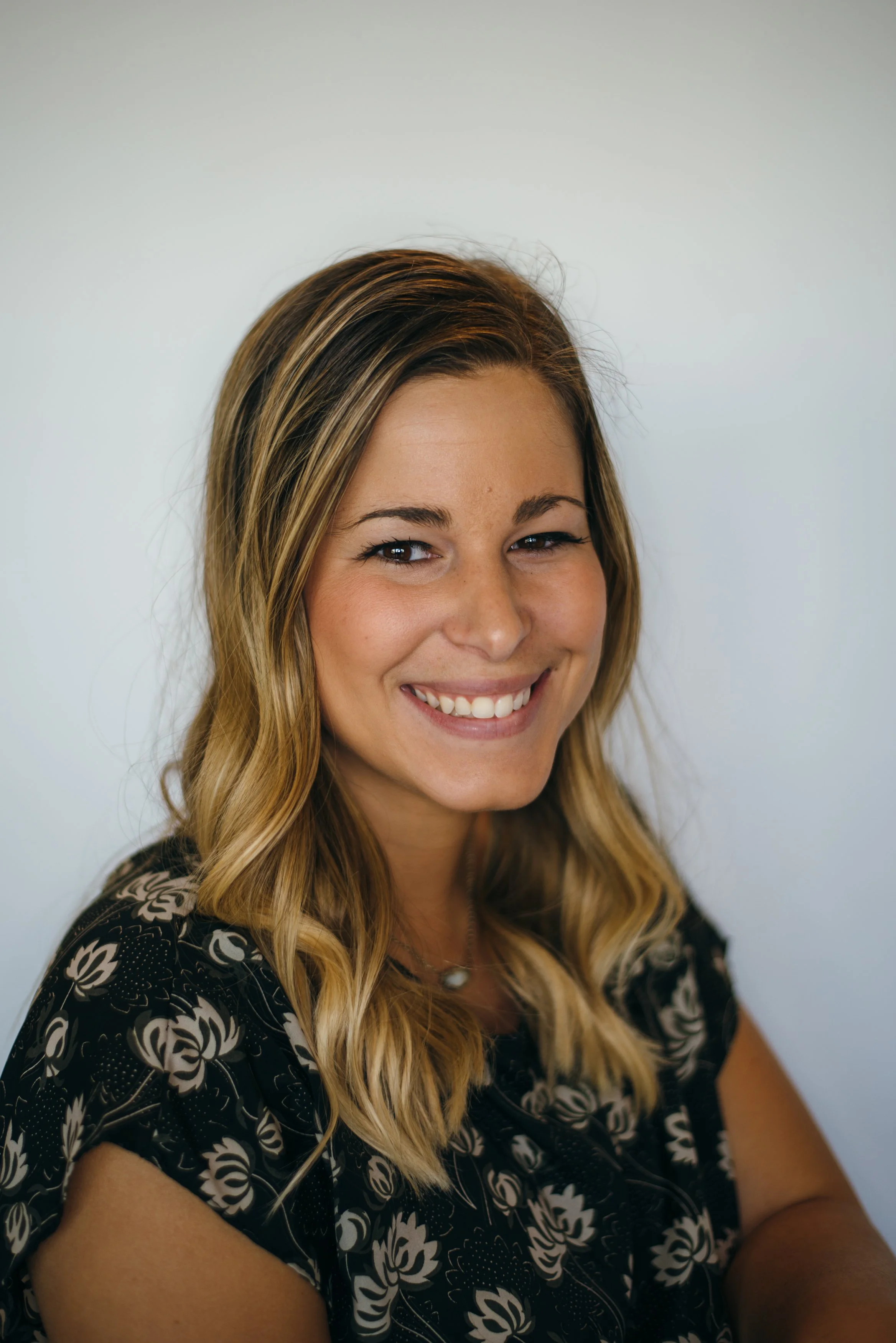 A young woman with long wavy blonde hair smiling at the camera, wearing a black top with a white floral pattern, against a plain light-colored background.