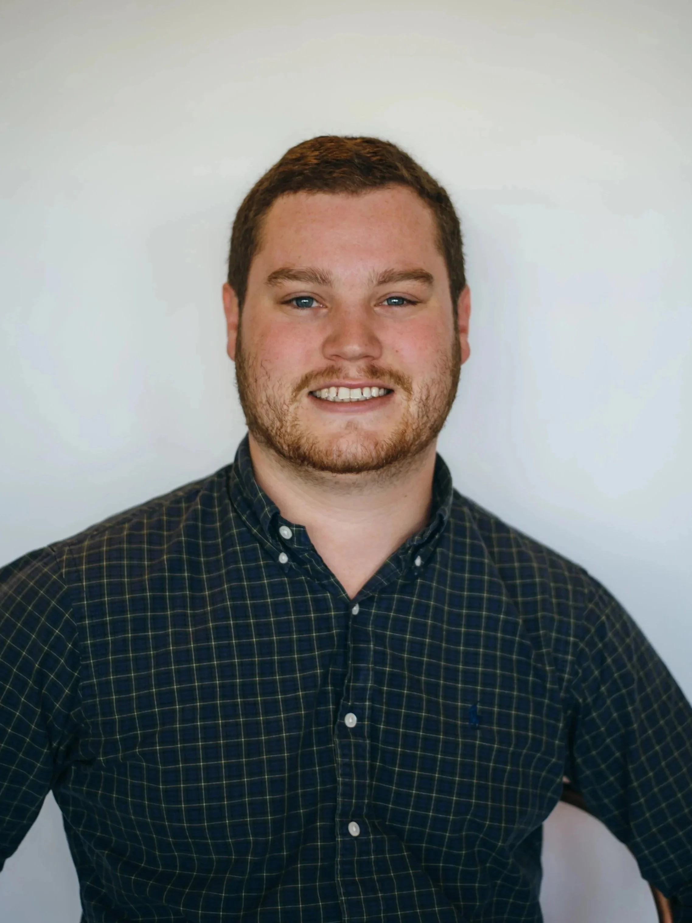 Close-up of a young man with short brown hair, wearing a dark plaid button-up shirt, smiling against a plain white background.