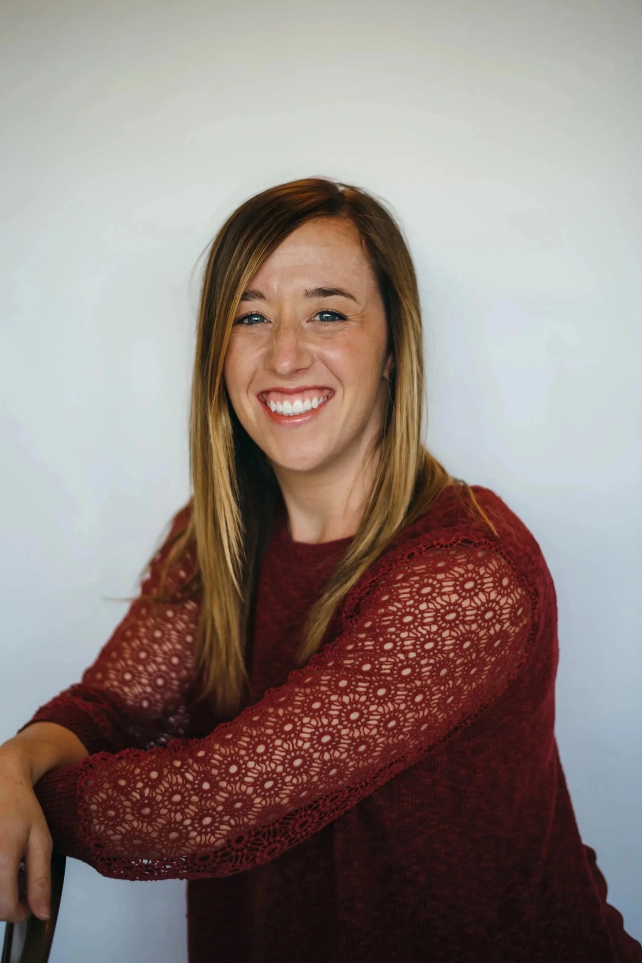 A woman with long, straight, light brown hair, smiling with her teeth visible, wearing a red lace long-sleeve top, sitting on a chair against a plain white wall.
