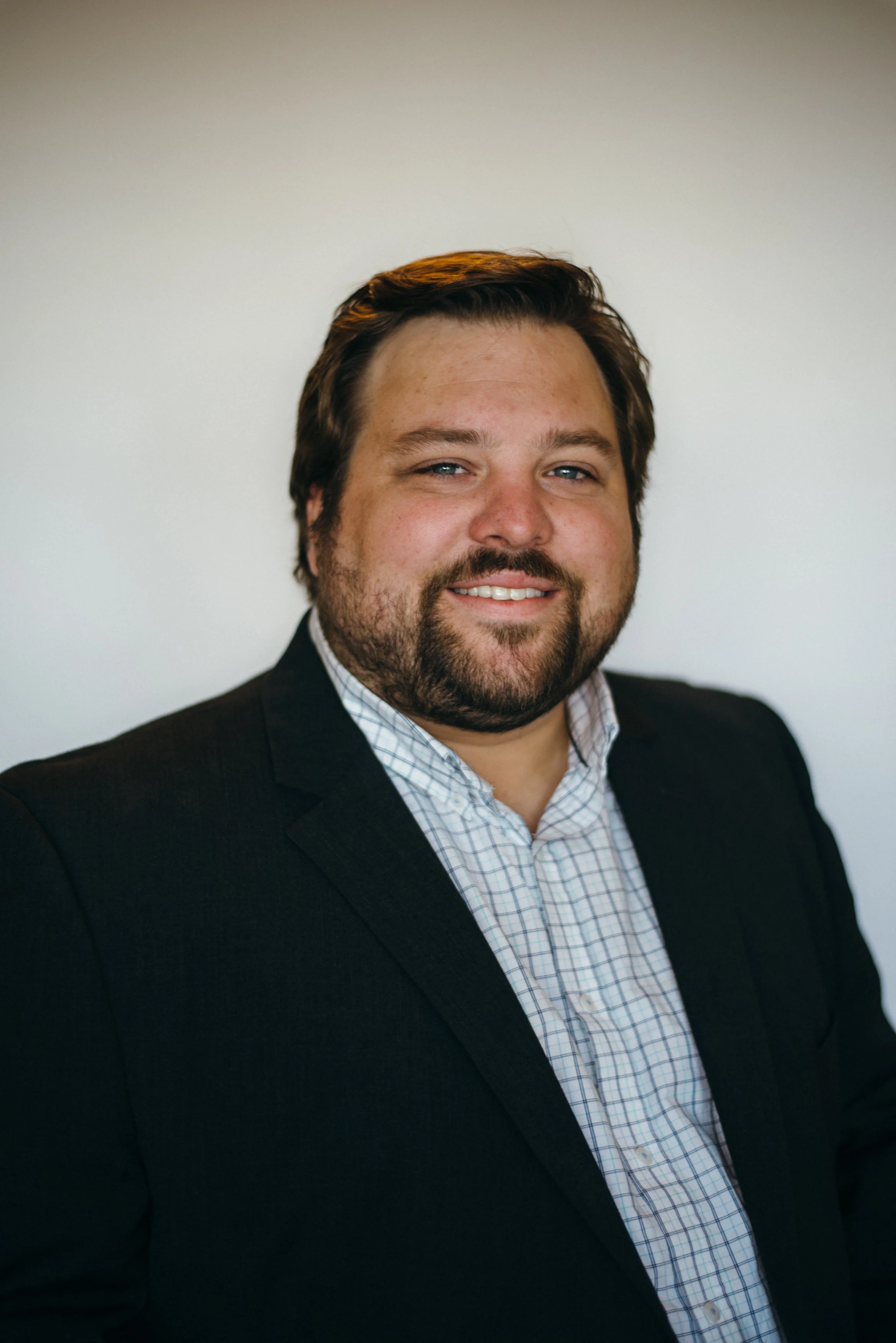 Headshot of a man with brown hair and beard, wearing a dark suit jacket and a light checkered shirt, smiling against a plain light background.