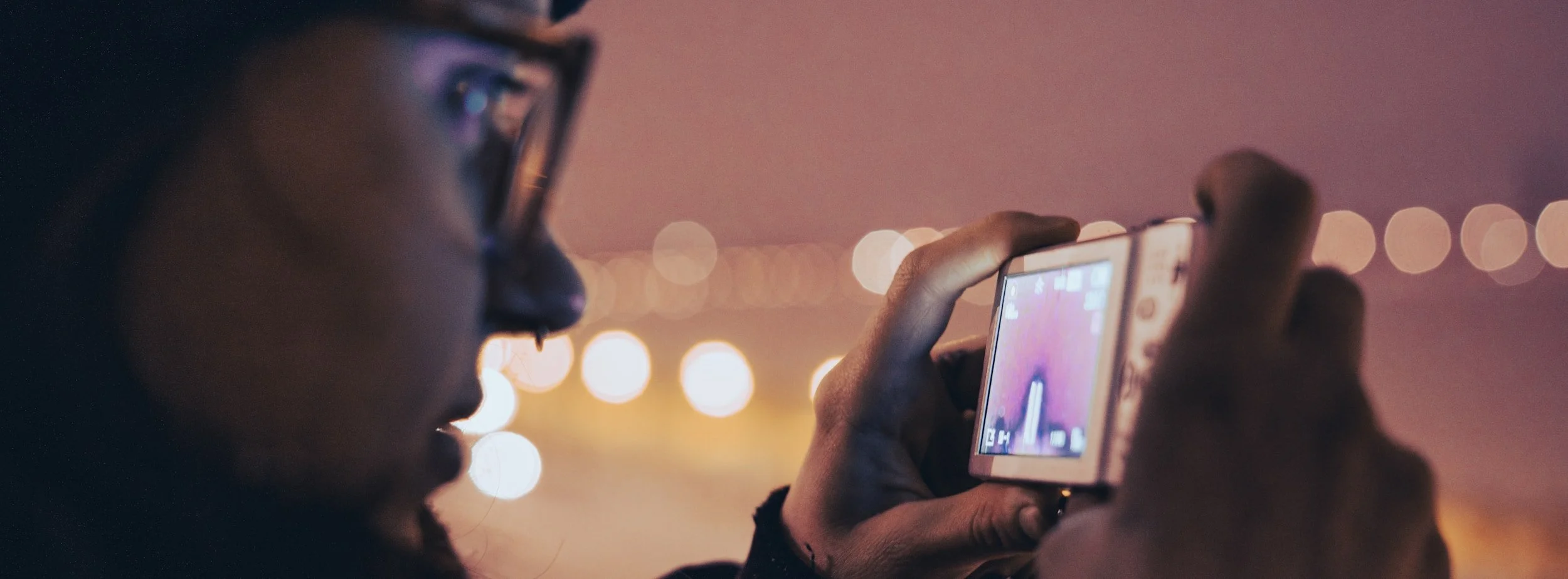 A person wearing glasses is taking a photo or recording with a digital camera or smartphone at night, with blurred city lights in the background.