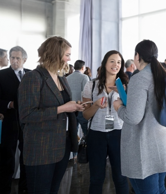 Three women are engaged in a conversation at a professional event, with other attendees mingling in the background in a bright conference room.