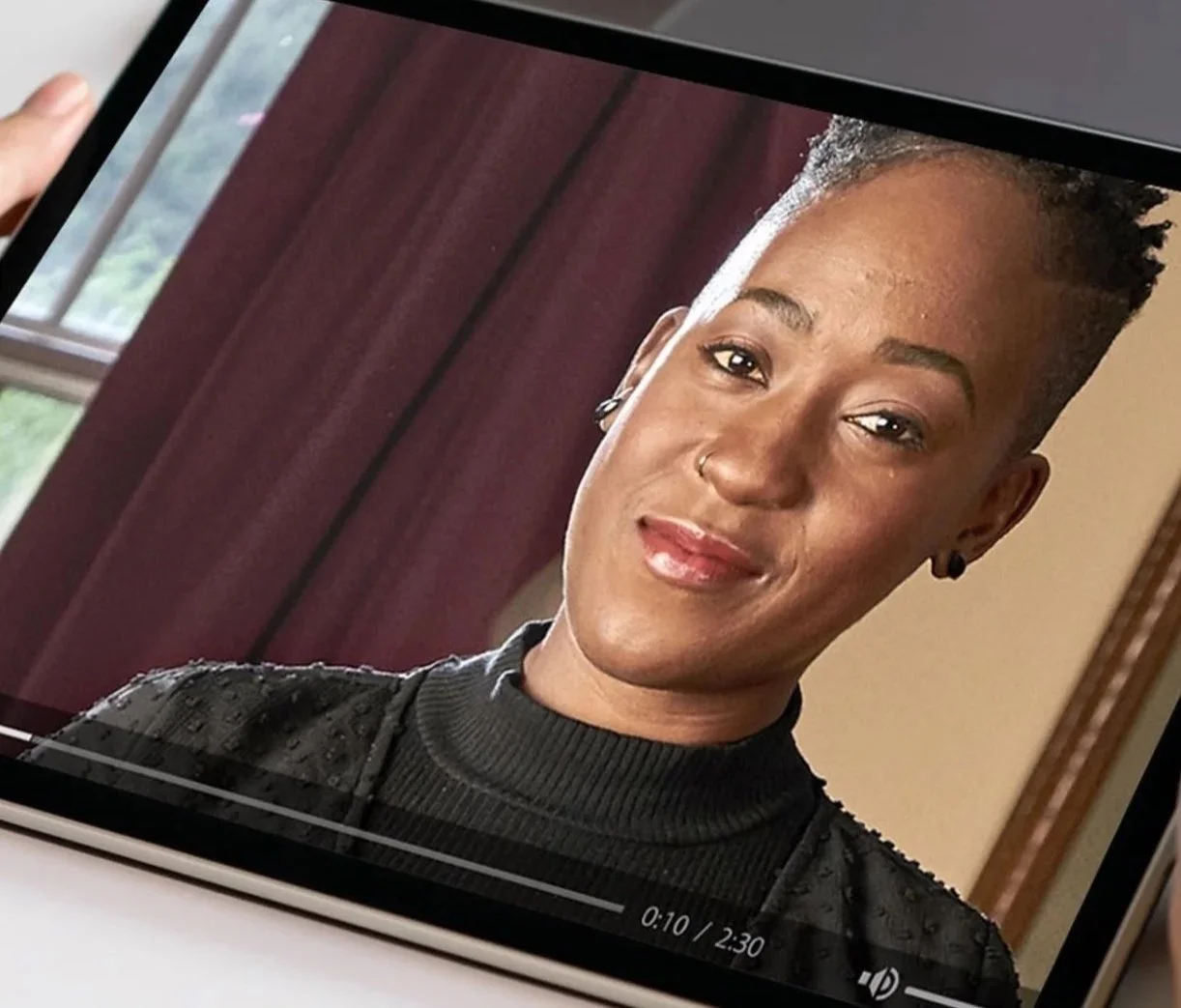 A woman with short, styled hair, wearing a black turtleneck sweater, smiling with earrings and a nose ring, in front of a wooden background.
