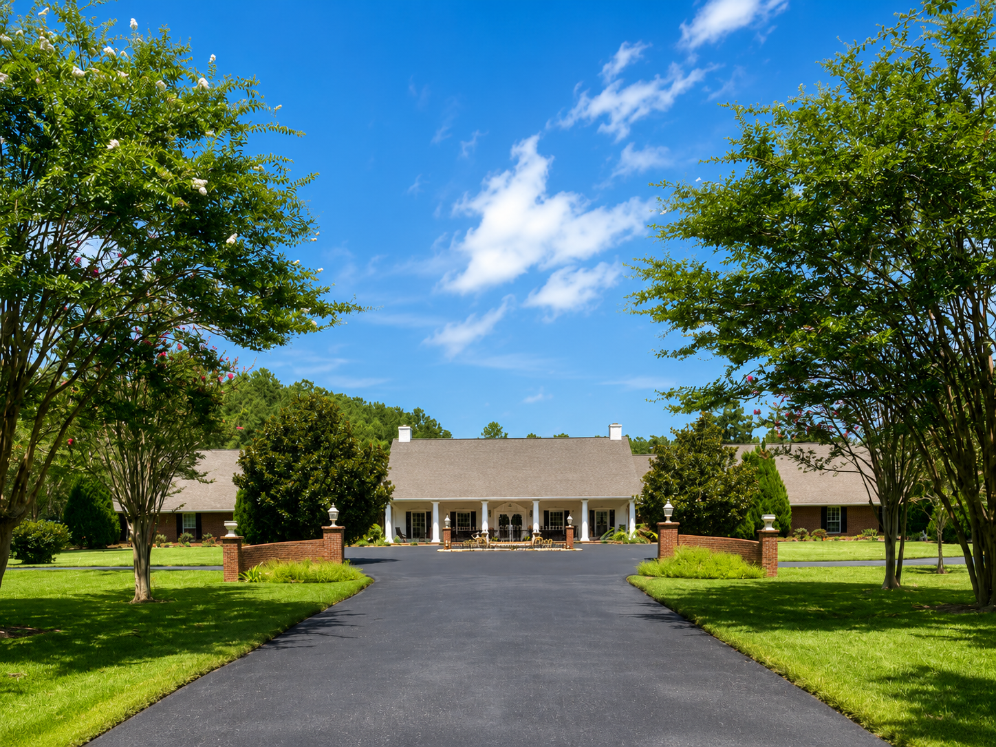 A large house with a covered porch, set back from a paved driveway, with a bright blue sky and scattered clouds above. The front yard features well-manicured grass, trees, and landscaped bushes.