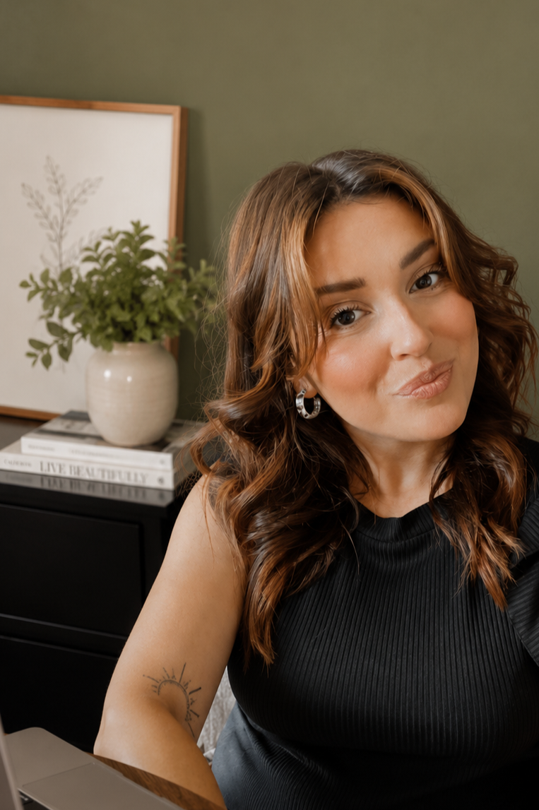 A woman with shoulder-length wavy brown hair smiling at the camera, sitting indoors near a desk with a potted plant, framed art, and books in the background.