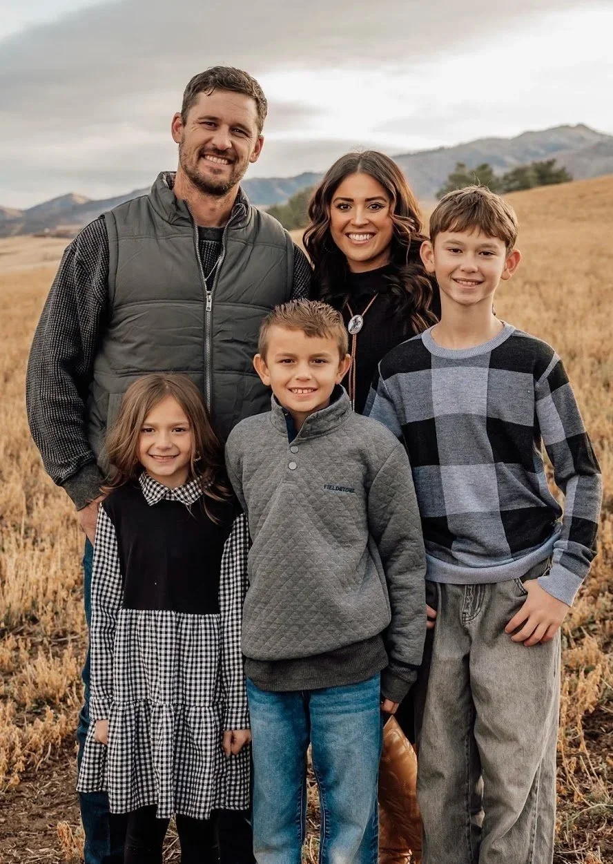A family of five stands outdoors in a field of dry grass with rolling hills in the background. The group includes two adult men and women and three children, all smiling and wearing casual clothing suitable for cool weather.