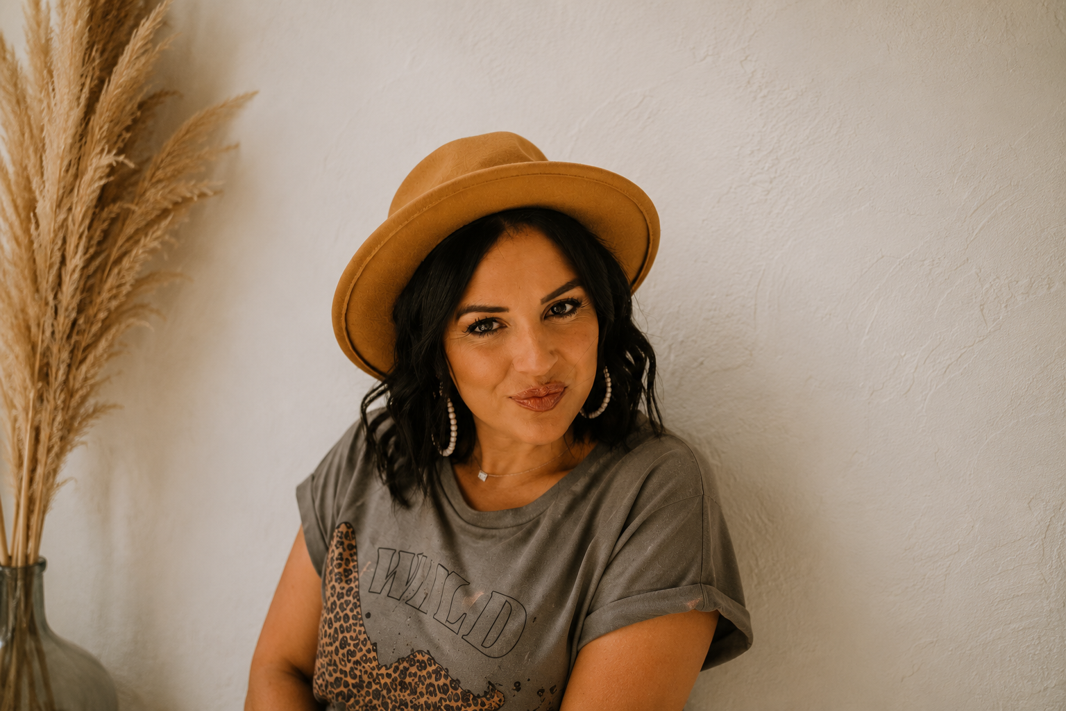 Woman wearing a tan wide-brim hat, gray graphic t-shirt, pearl earrings, and a necklace, smiling softly against a textured white wall with dried pampas grass in a vase.