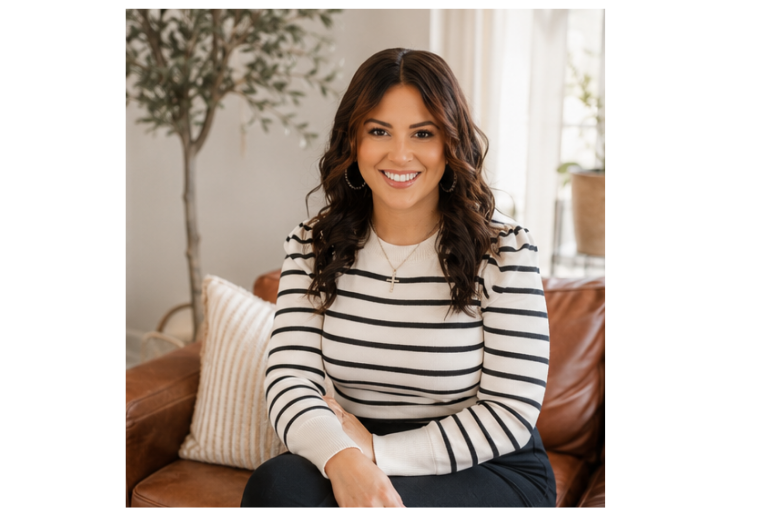 A woman with dark, curly hair smiling and sitting on a brown leather couch with cream-colored pillows in a well-lit room with a window and a potted plant in the background.