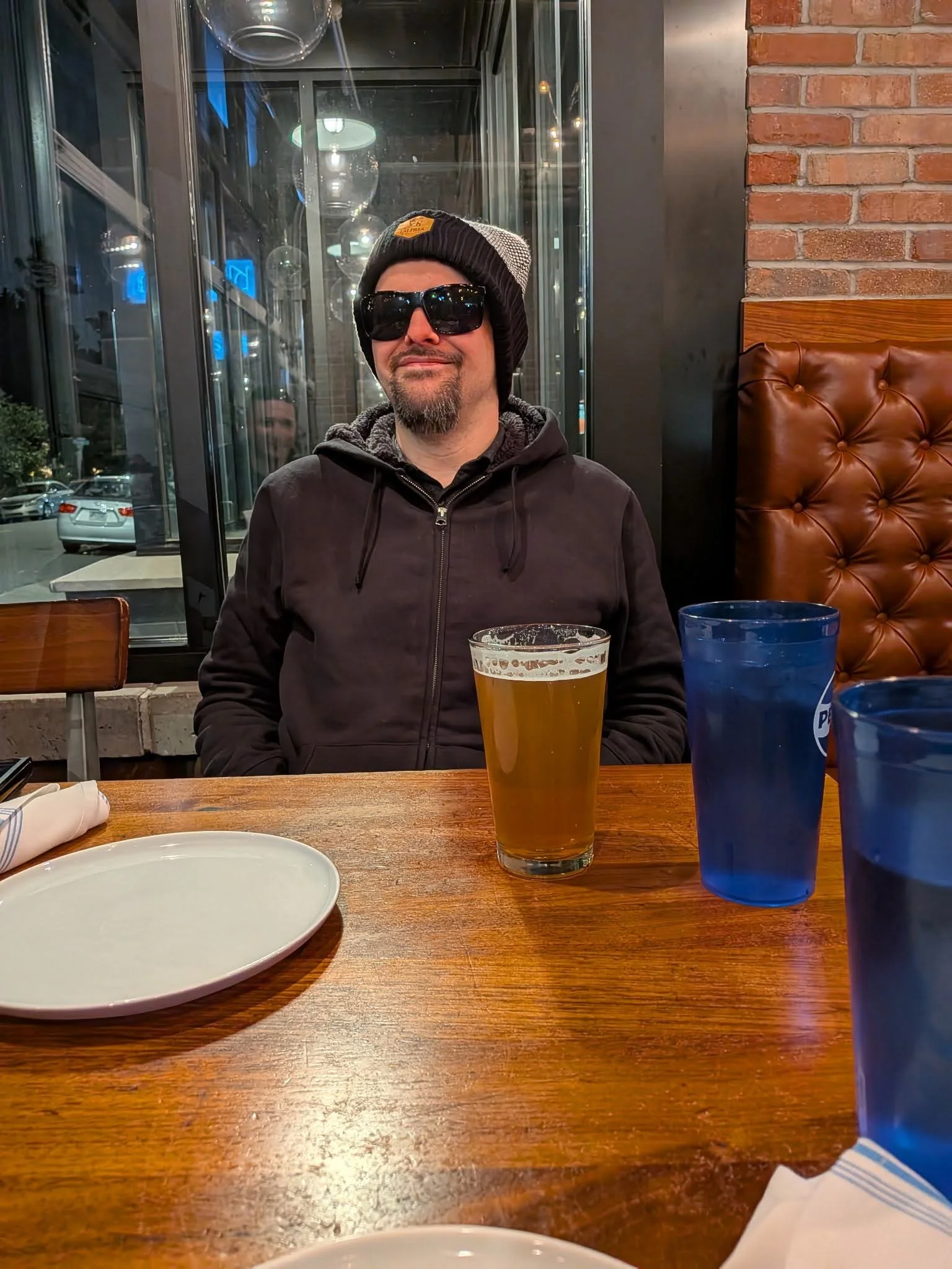 A man sitting at a restaurant table wearing a black hoodie, black beanie, and sunglasses, with a beard and mustache. On the table are three drinks in tall glasses and a white plate. The background shows a brick wall and large windows with reflections.
