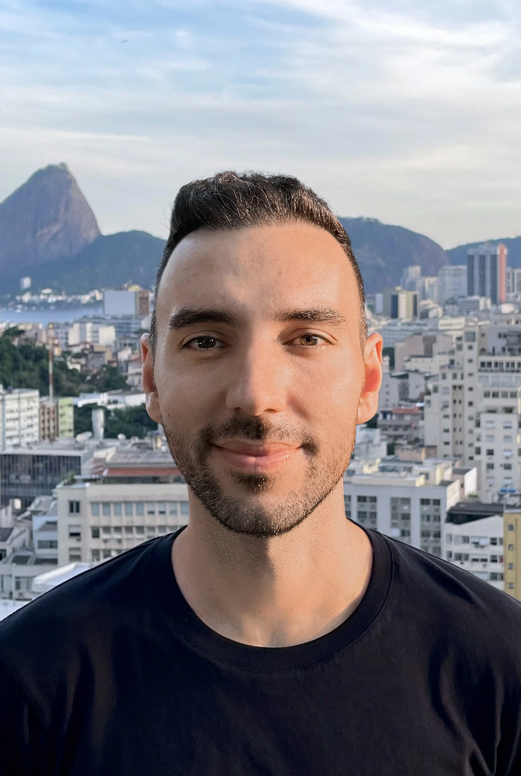 Close-up of a smiling young man with short dark hair and facial hair, wearing a black T-shirt, standing on a rooftop with a city skyline and mountains in the background during daylight.