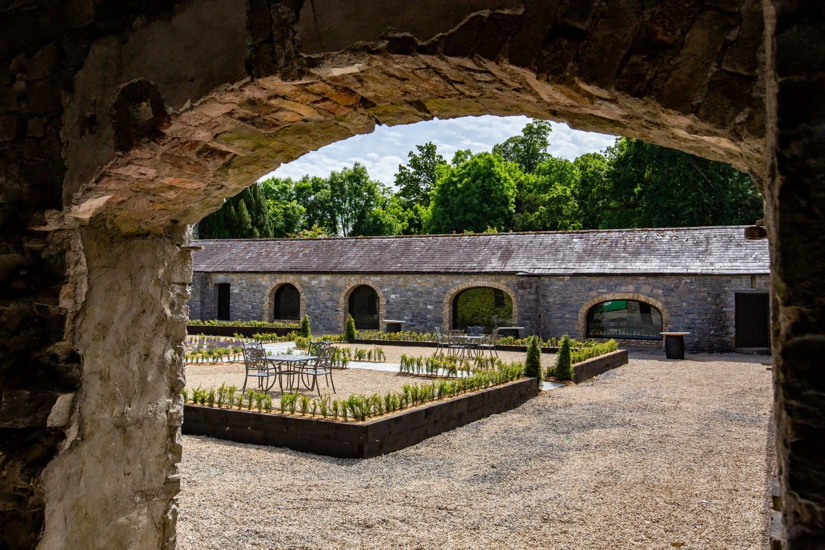 View of a courtyard with stone building and garden tables, framed by an archway made of stone bricks, with trees and a cloudy sky in the background.