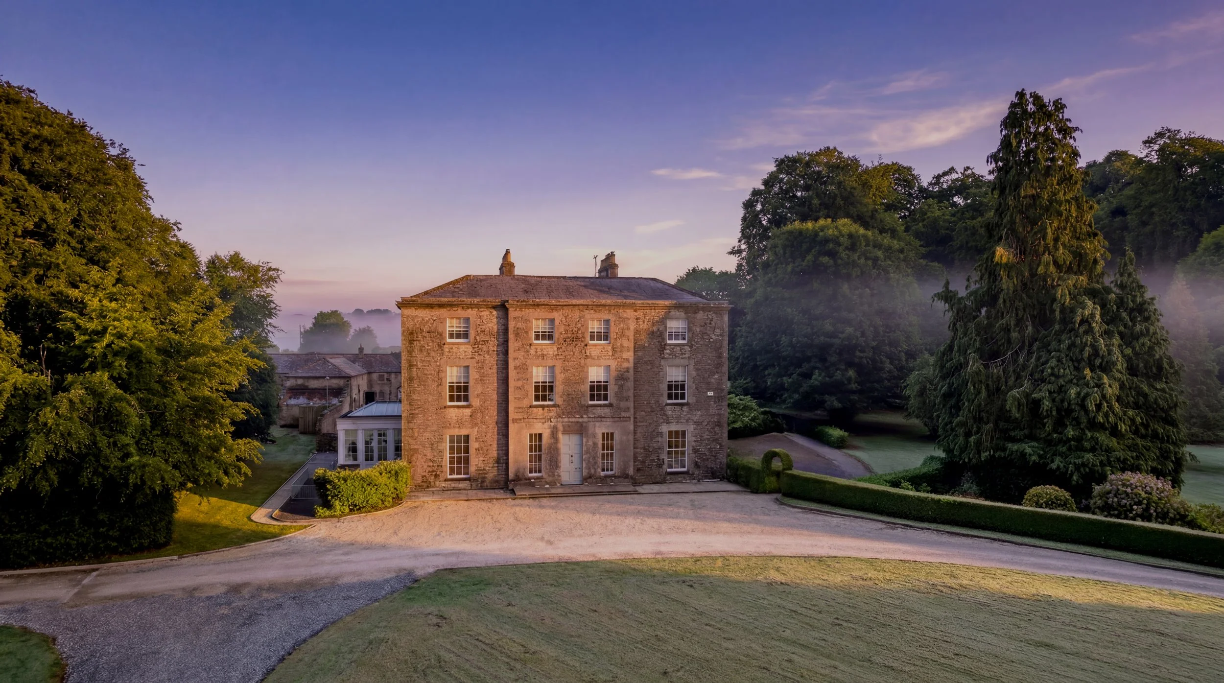 A large stone house surrounded by well-manicured grass and trees, with a gravel driveway in the foreground and a peaceful, slightly misty landscape under a purple and blue sky.