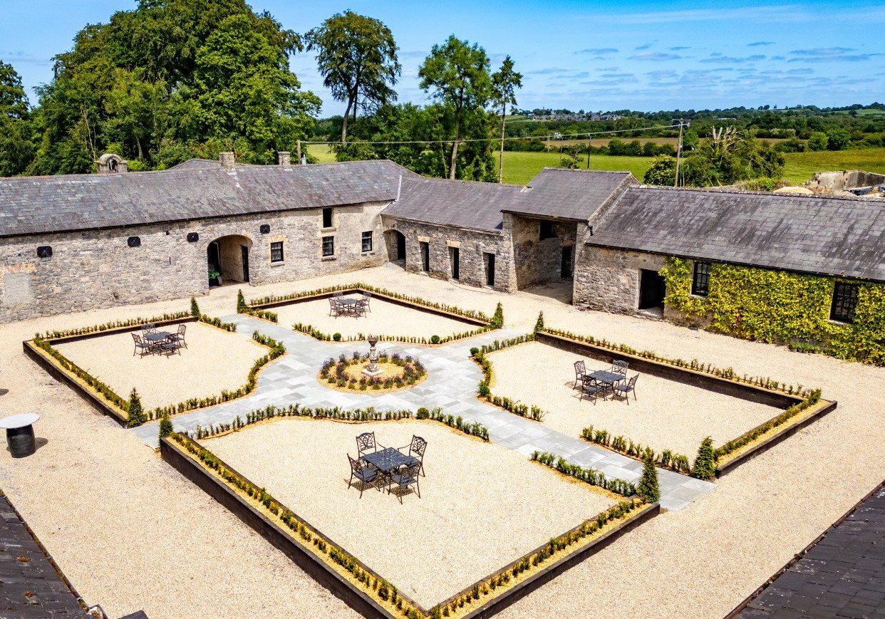 A courtyard with four sections of gravel, each with wrought iron tables and chairs, surrounded by low yellow and green hedge borders, with a central fountain and paved walkways connecting the sections, all enclosed by stone buildings and green trees in the background.