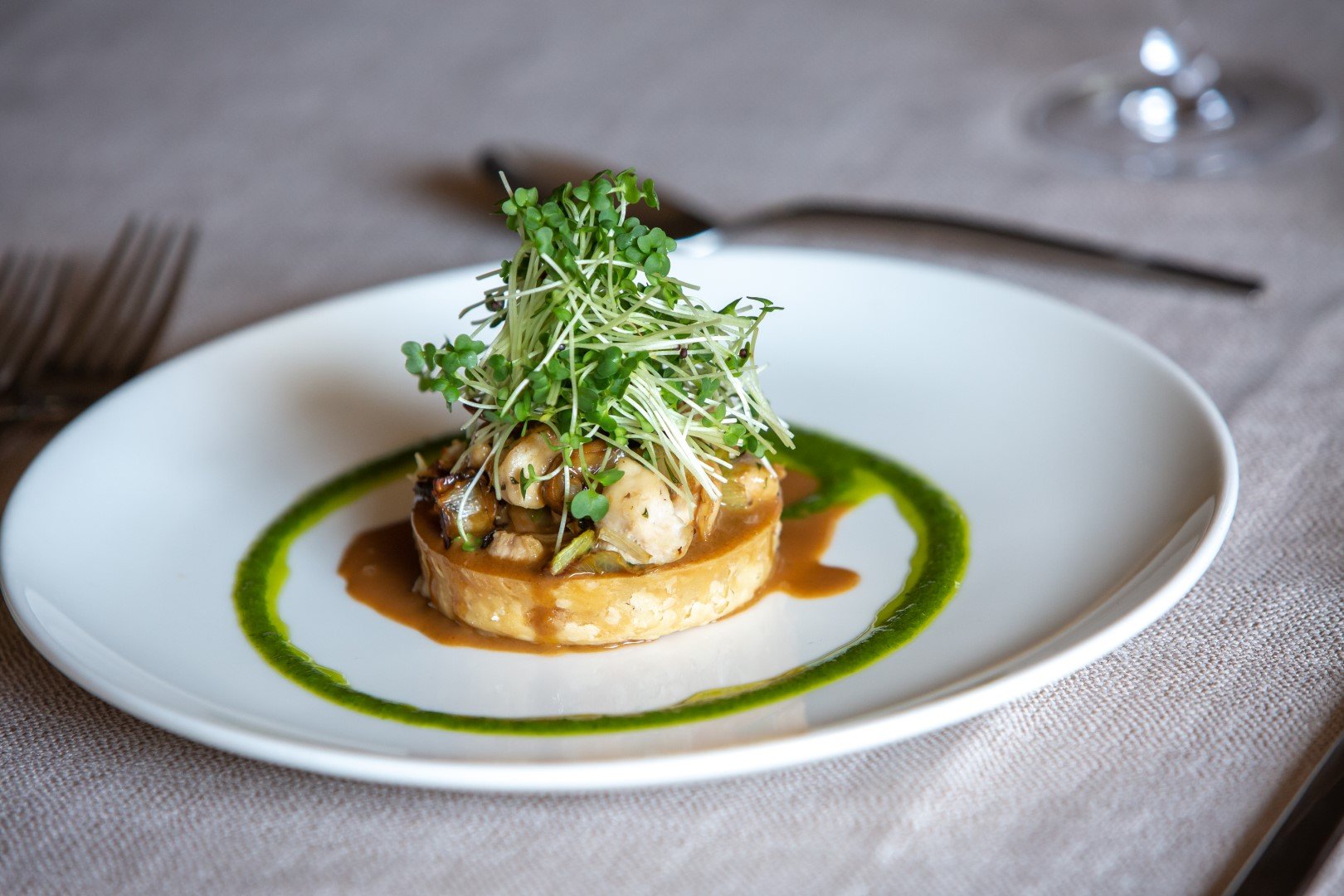 A gourmet appetizer featuring a round base topped with cooked seafood and garnished with microgreens, served on a white plate with a green sauce accent.