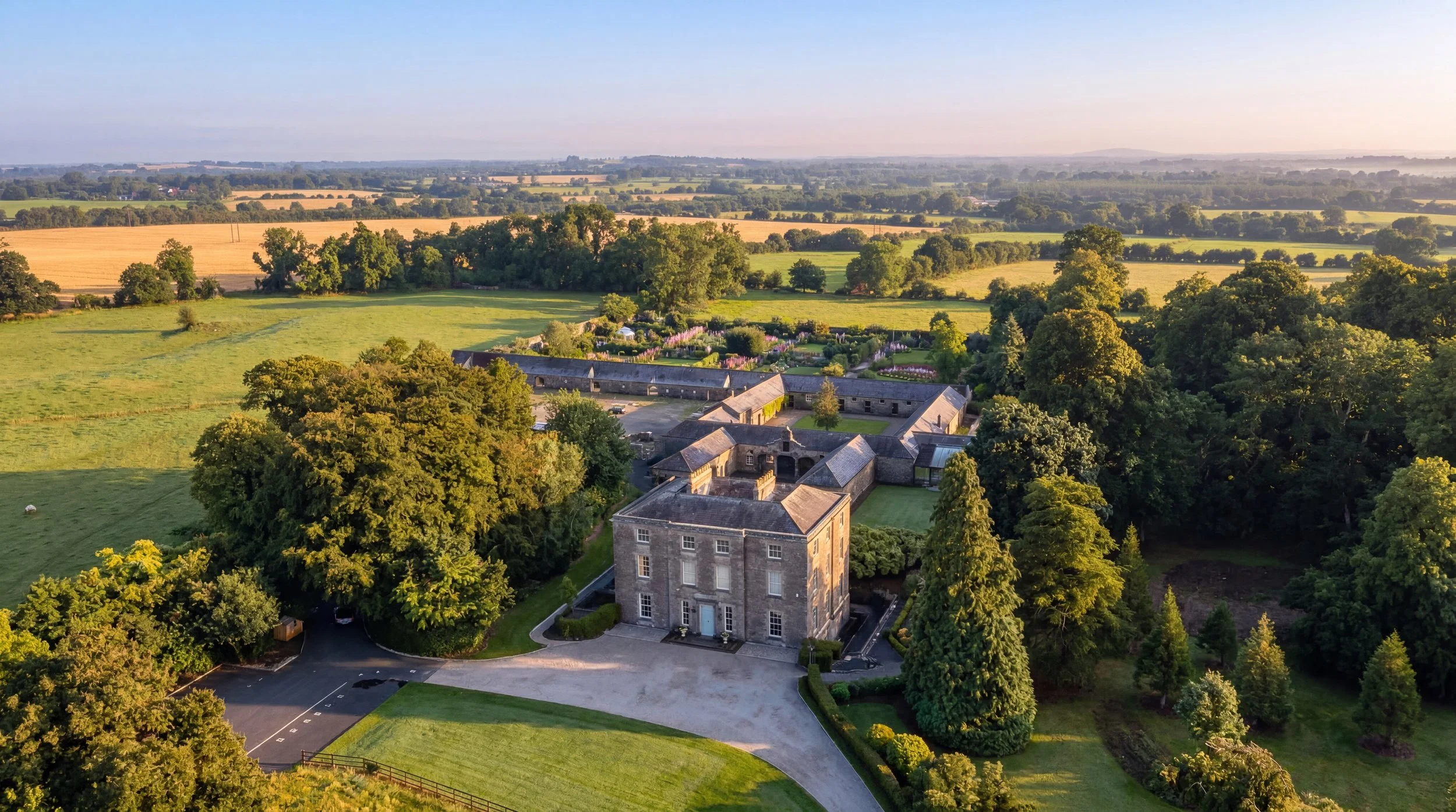 Aerial view of an estate with a large stone mansion surrounded by lush trees and gardens, expansive fields, and rolling countryside under a clear sky.