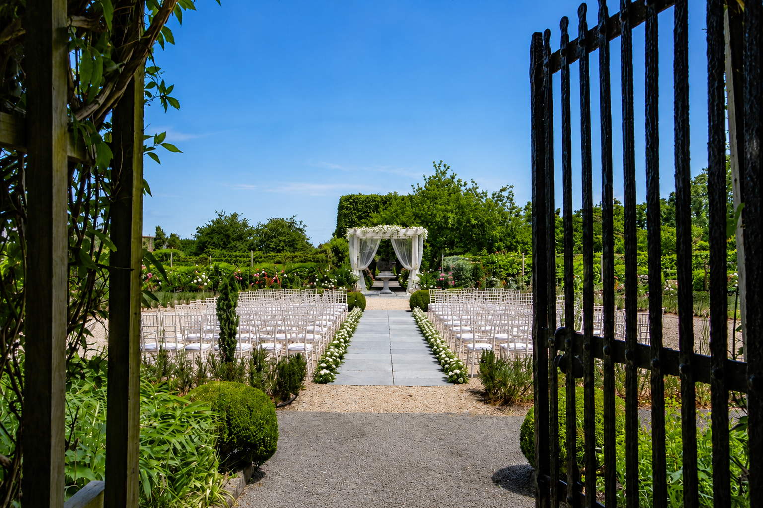 View of an outdoor wedding ceremony setup with white chairs arranged on both sides of a stone aisle leading to a white canopy with sheer curtains, surrounded by green plants and trees under a clear blue sky.