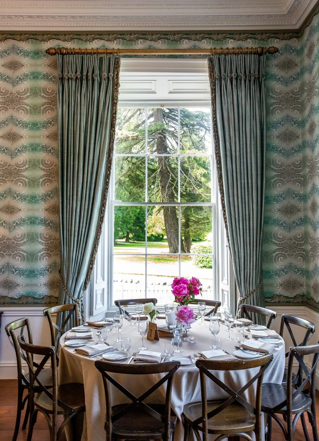 Round dining table set for a formal meal with a white tablecloth, crystal glasses, white napkins, and floral centerpieces with pink and white flowers, situated by large window with trees outside and decorated curtains.
