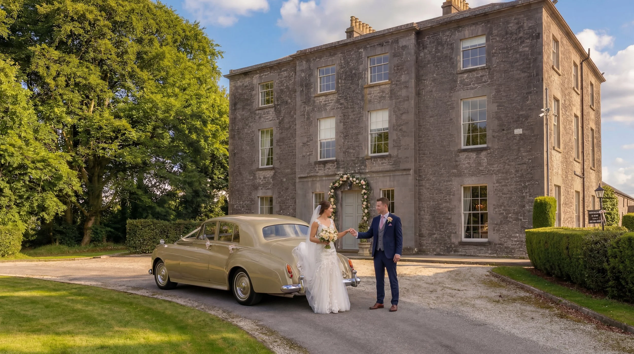 A bride and groom stand outside a stone building decorated with floral wedding decorations, holding hands and smiling at each other, with a vintage car nearby.