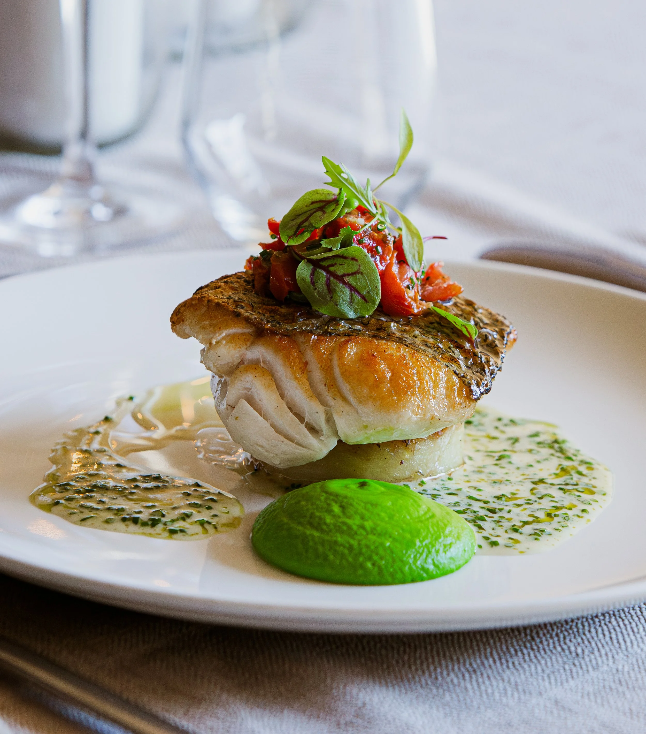 A gourmet dish featuring a piece of grilled fish topped with tomato relish and microgreens, served on a white plate with green sauce and garnished with lime slices, on a table set with empty wine glasses.