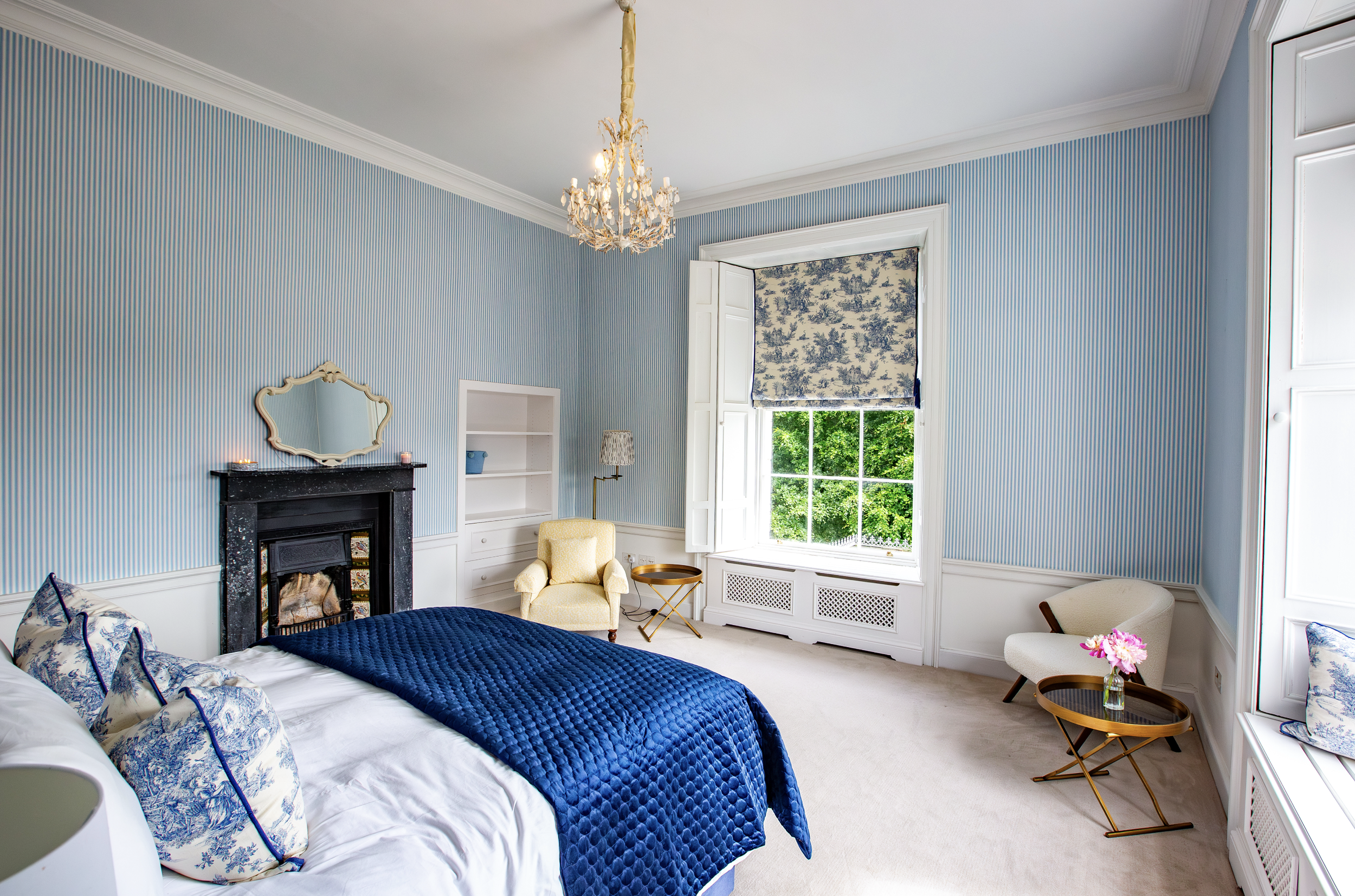 A bright bedroom with light blue and white striped wallpaper, a large window with a decorative Roman shade, a black fireplace, a yellow armchair, a white built-in bookshelf, a table with a pink flower, and a chandelier hanging from the ceiling.