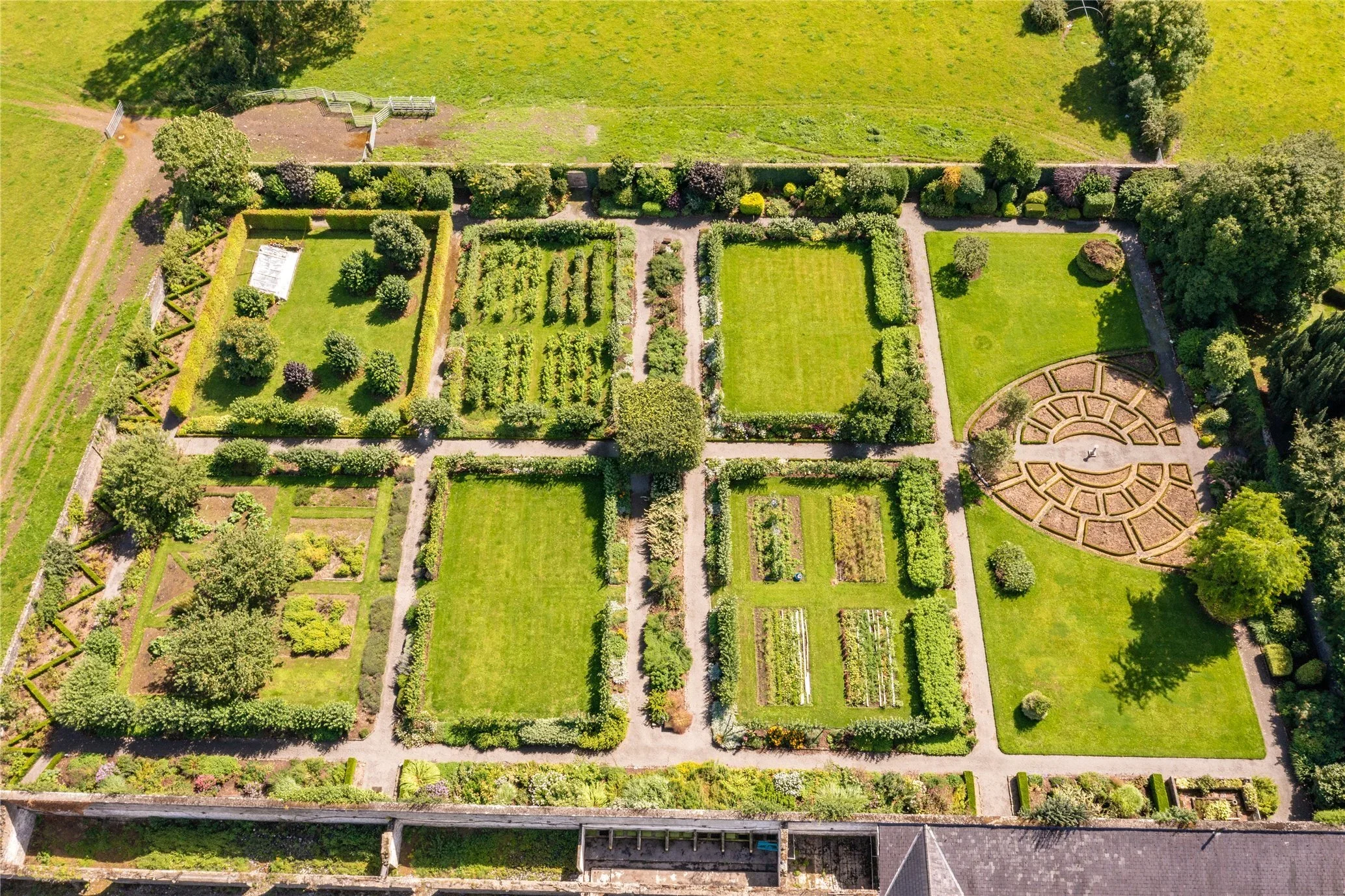 An aerial view of a large, well-maintained garden with various plots, pathways, trees, and a circular patterned area, adjacent to grassy fields.