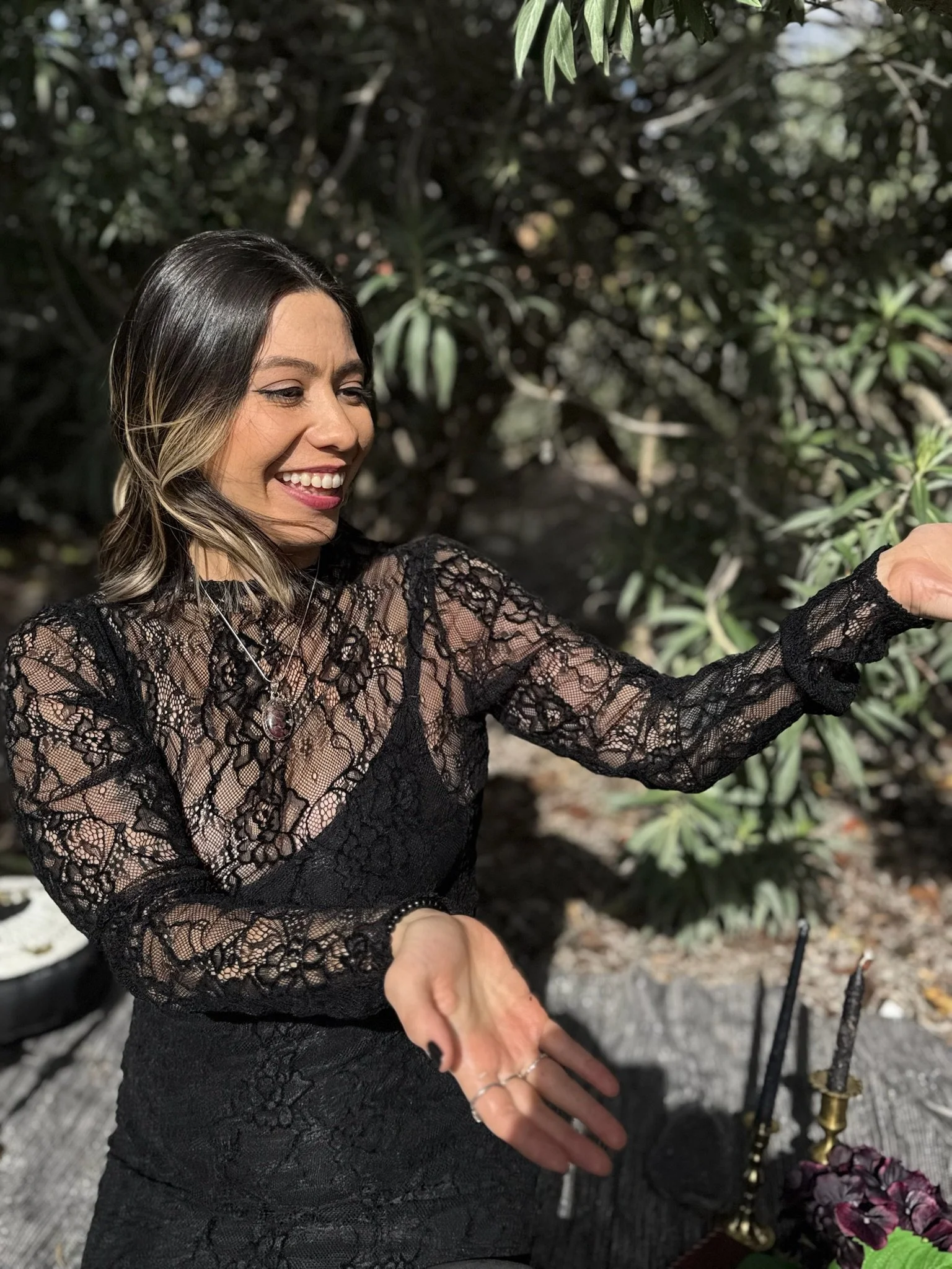 A woman with shoulder-length dark hair styled with blonde highlights and light makeup, wearing a black lace top and multiple rings, is smiling and taking a selfie outdoors among green foliage.