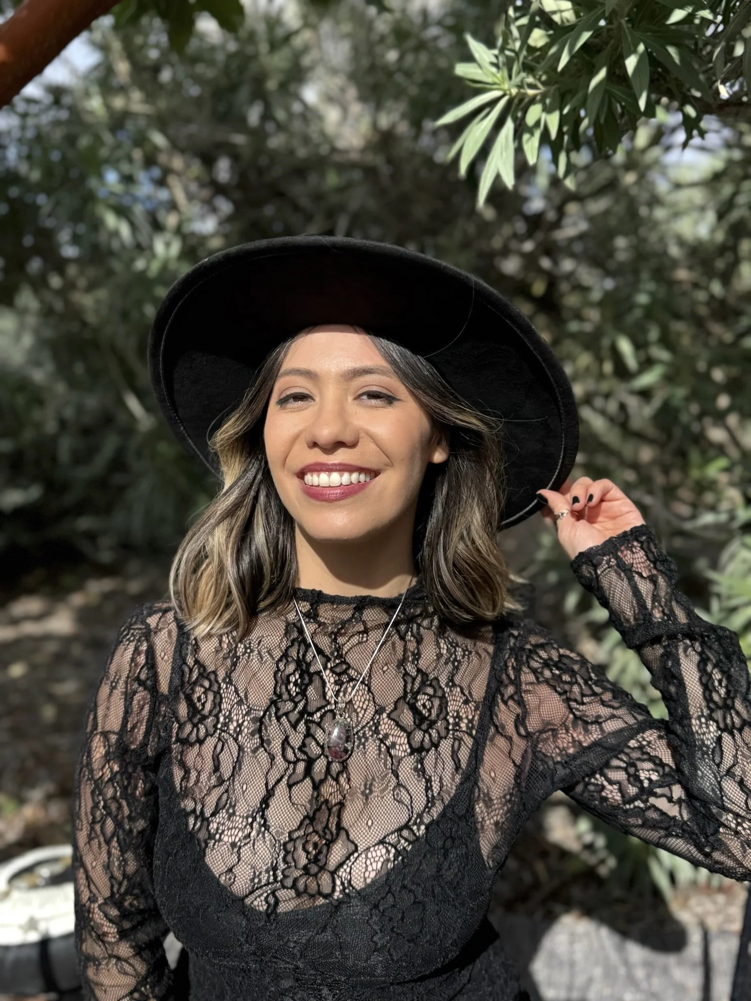 A woman smiling and wearing a black wide-brimmed hat and a black lace dress with jewelry, outdoors with greenery in the background.