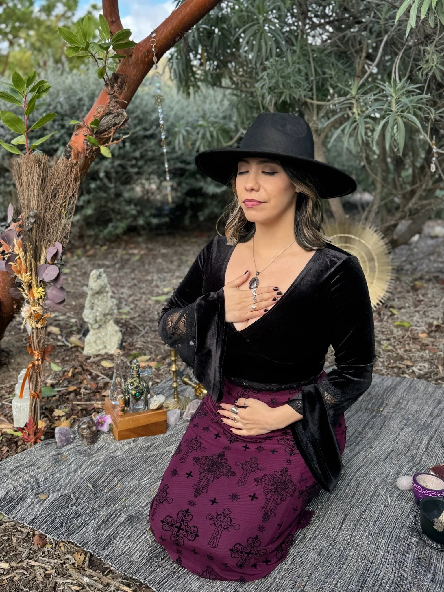 A woman in a black hat and black velvet jacket with a purple skirt featuring symbols, sitting on a rug outdoors with eyes closed and hand over heart, surrounded by spiritual objects and greenery.